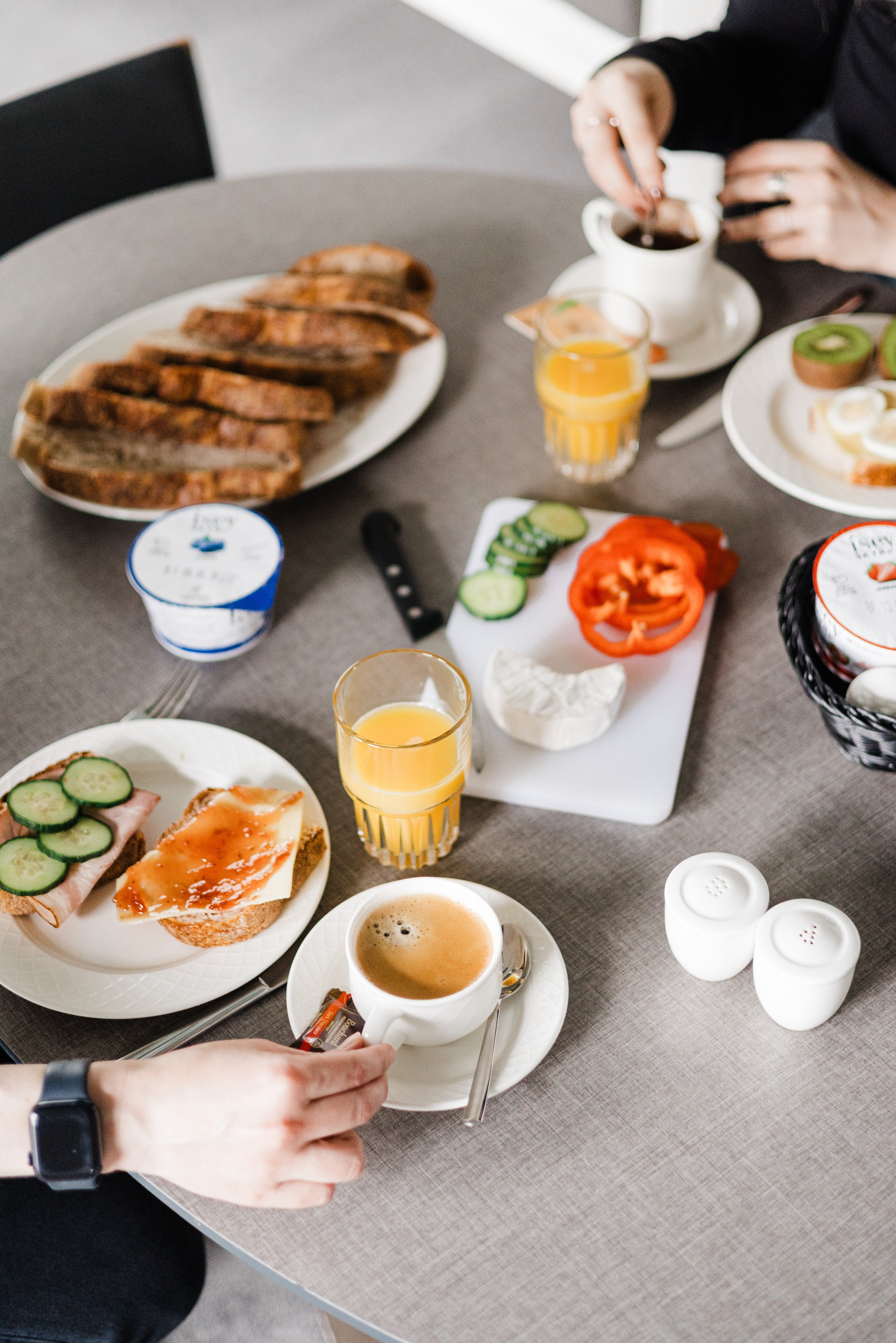 Breakfast spread on a table including sliced toast, cucumber slices with ham, jam, cream cheese, sliced red pepper, poached egg on toast, glasses of orange juice, a cup of coffee, and some yogurt containers.