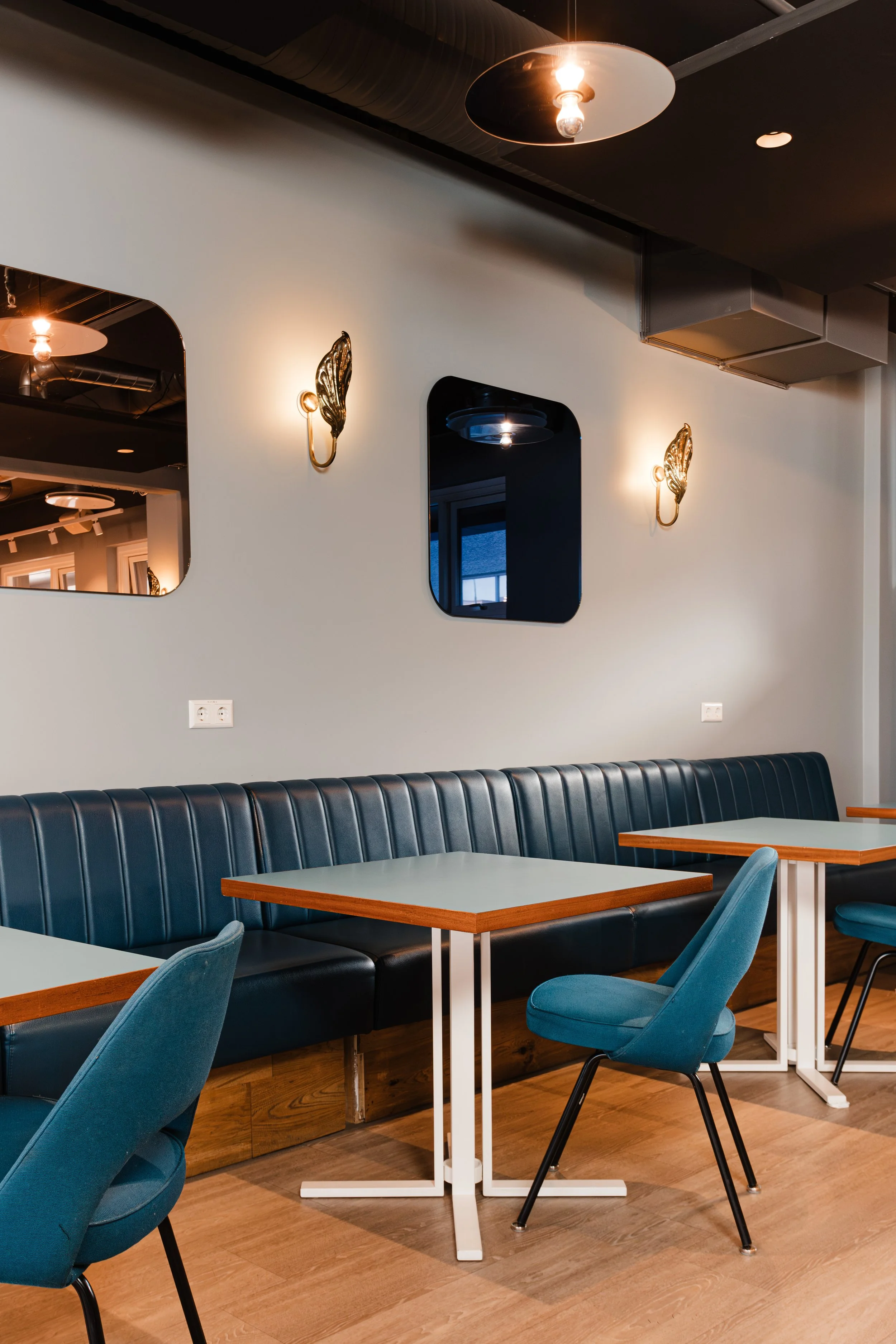 Empty modern restaurant with blue chairs, black leather bench seating, light wood tables, mirrors, and decorative wall sconces.