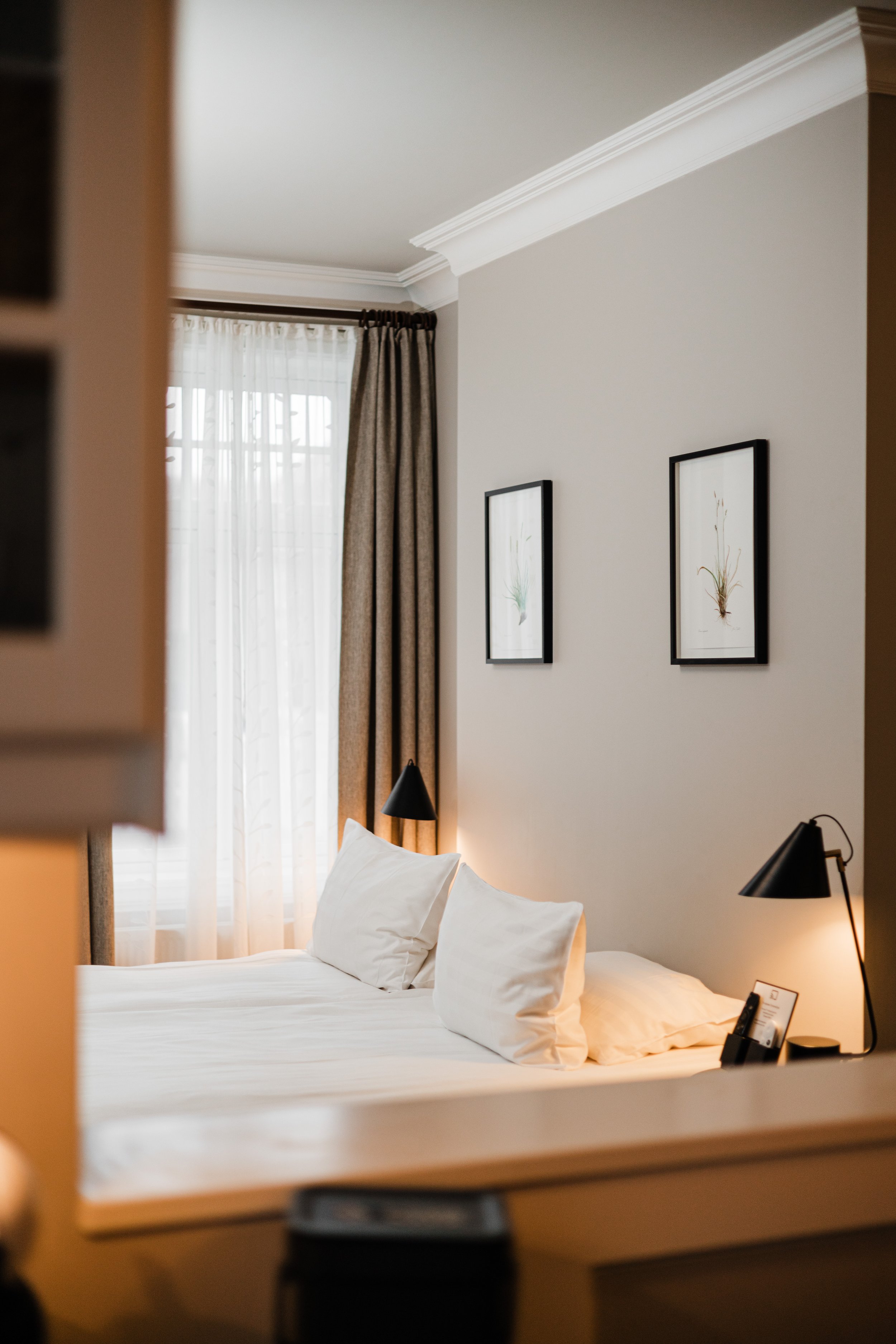 A cozy hotel bedroom with a neatly made bed, white pillows, framed botanical art on the wall, a window with sheer and blackout curtains, and two black bedside lamps.
