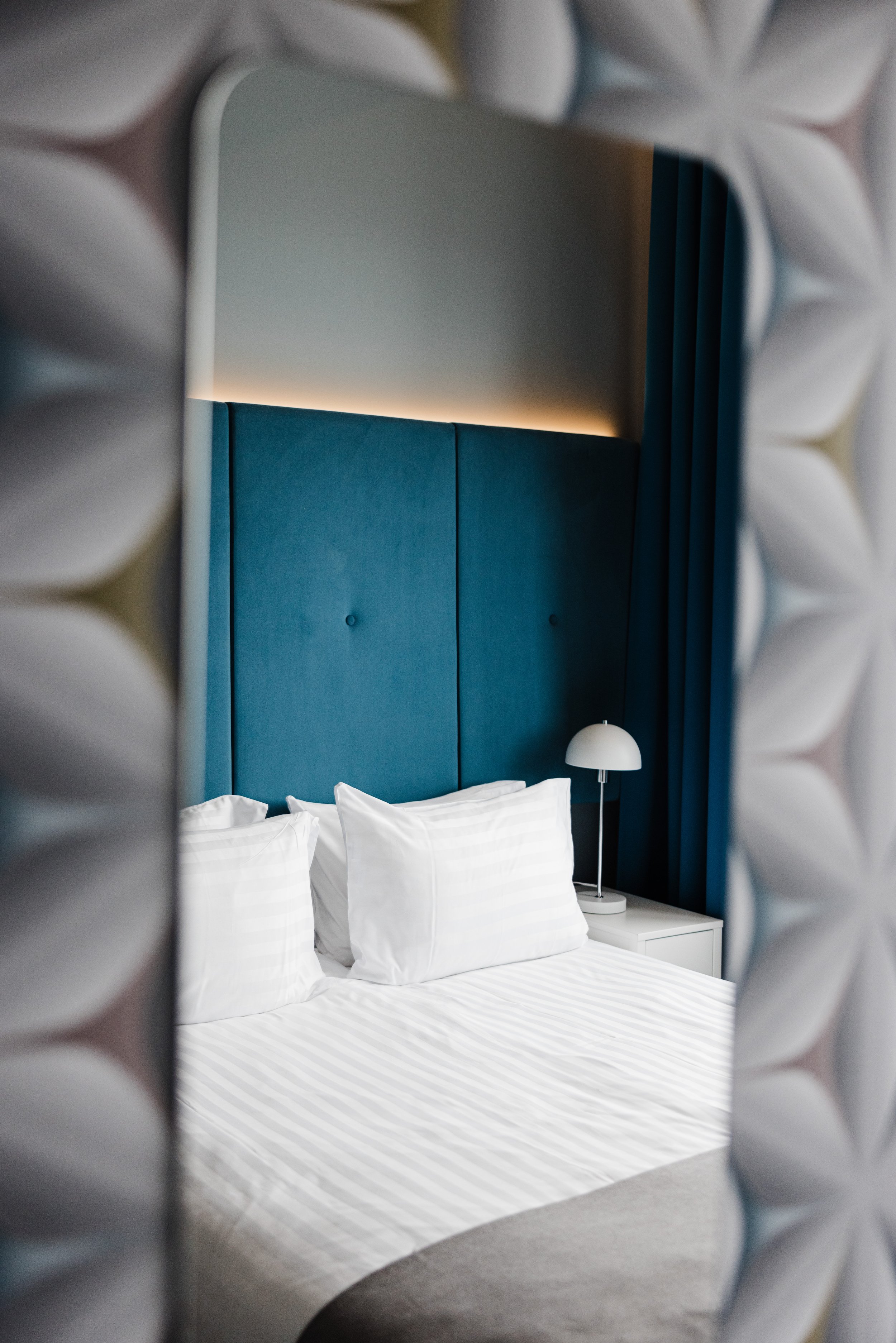 A hotel room viewed through a decorative mirror with petal-shaped frame, showing a bed with white striped bedding, white pillows, a blue upholstered headboard, a white bedside table, and a white modern table lamp, with blue curtains in the background.