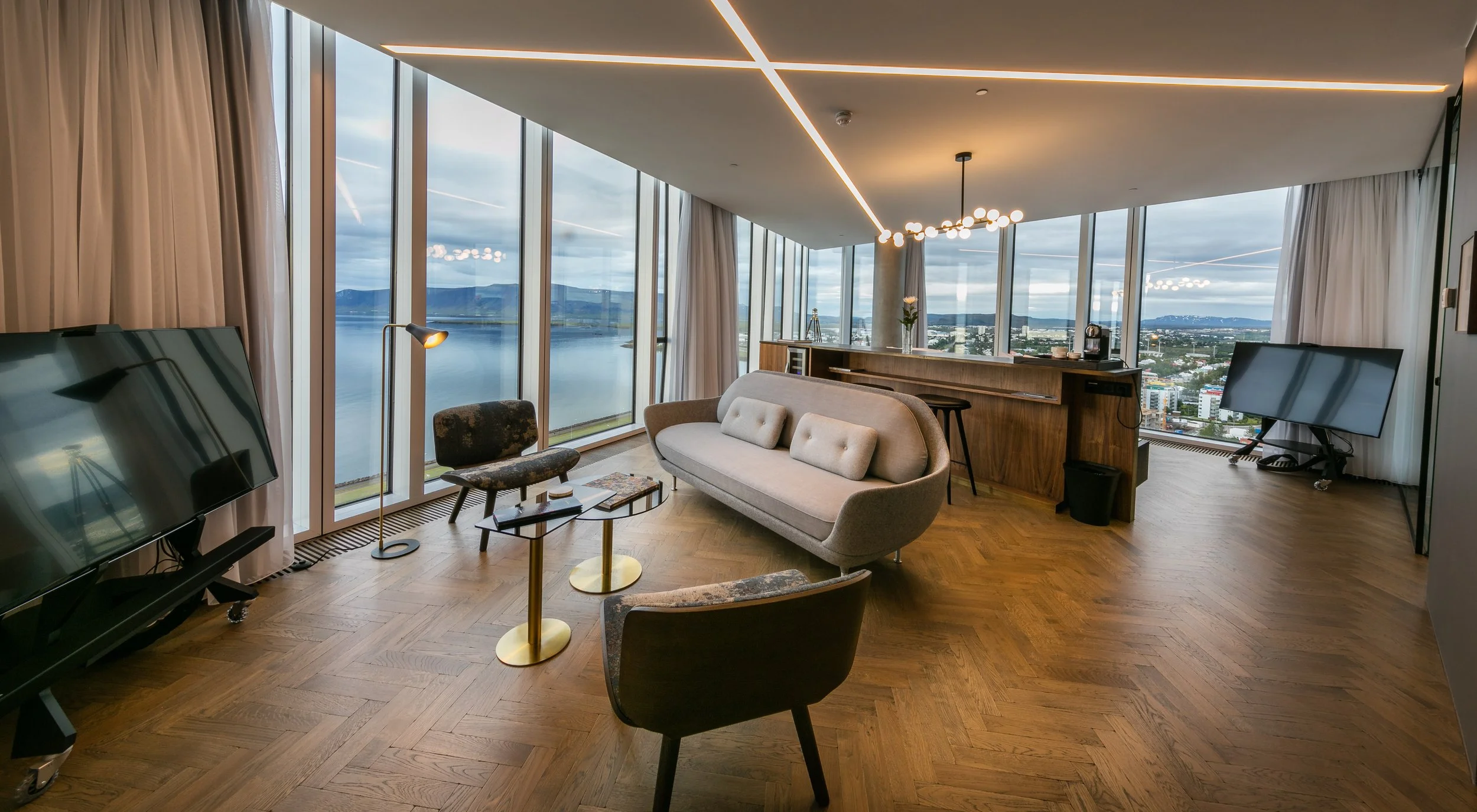 Modern hotel living room with large floor-to-ceiling windows showing water and city view, featuring a gray curved sofa, two patterned chairs, a small glass coffee table, a television on a stand, and a bar area with stools, all with wood flooring and minimalist decor.