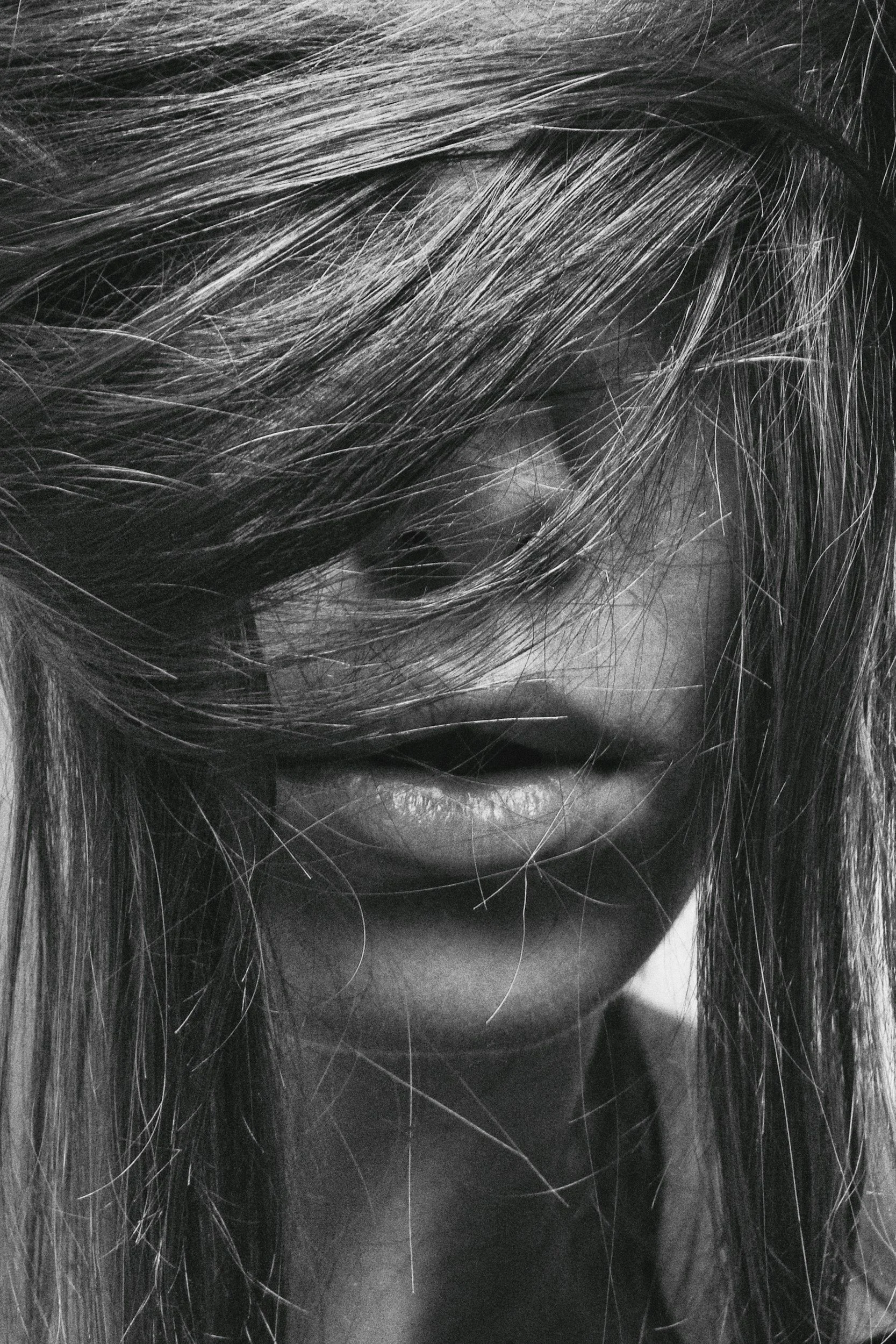Close-up black and white photograph of a woman's face mostly obscured by her messy hair, with only her lips and part of her nose visible.