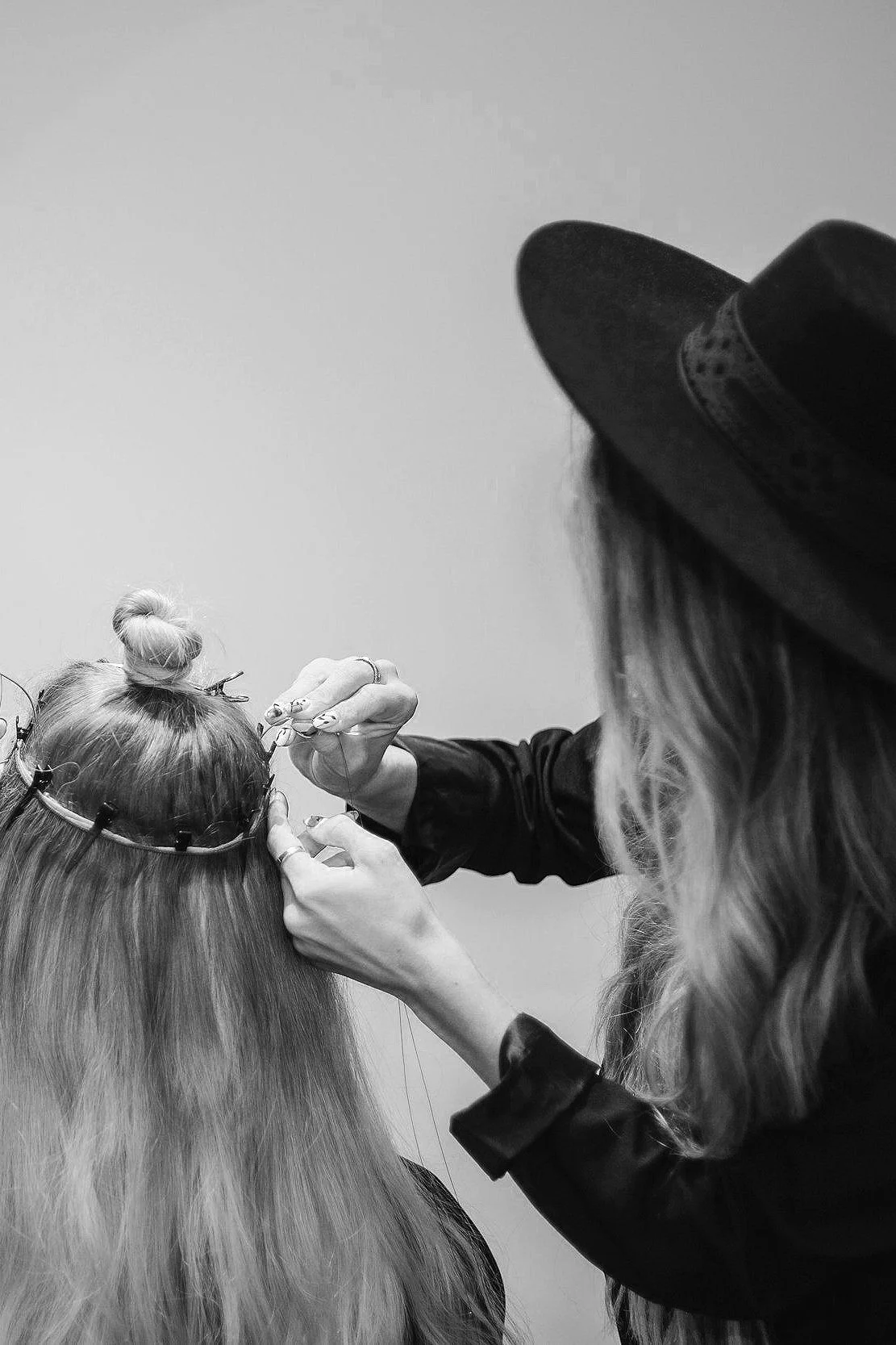 A woman with long hair wearing a hat is helping another woman with long hair to put a floral crown on her head.