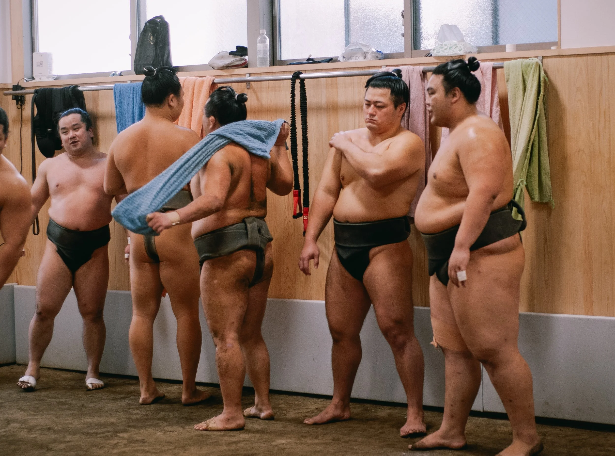 A group of sumo wrestlers in a training facility, dressed in traditional mawashi, standing on a mat with towels and water bottles on a shelf behind them.