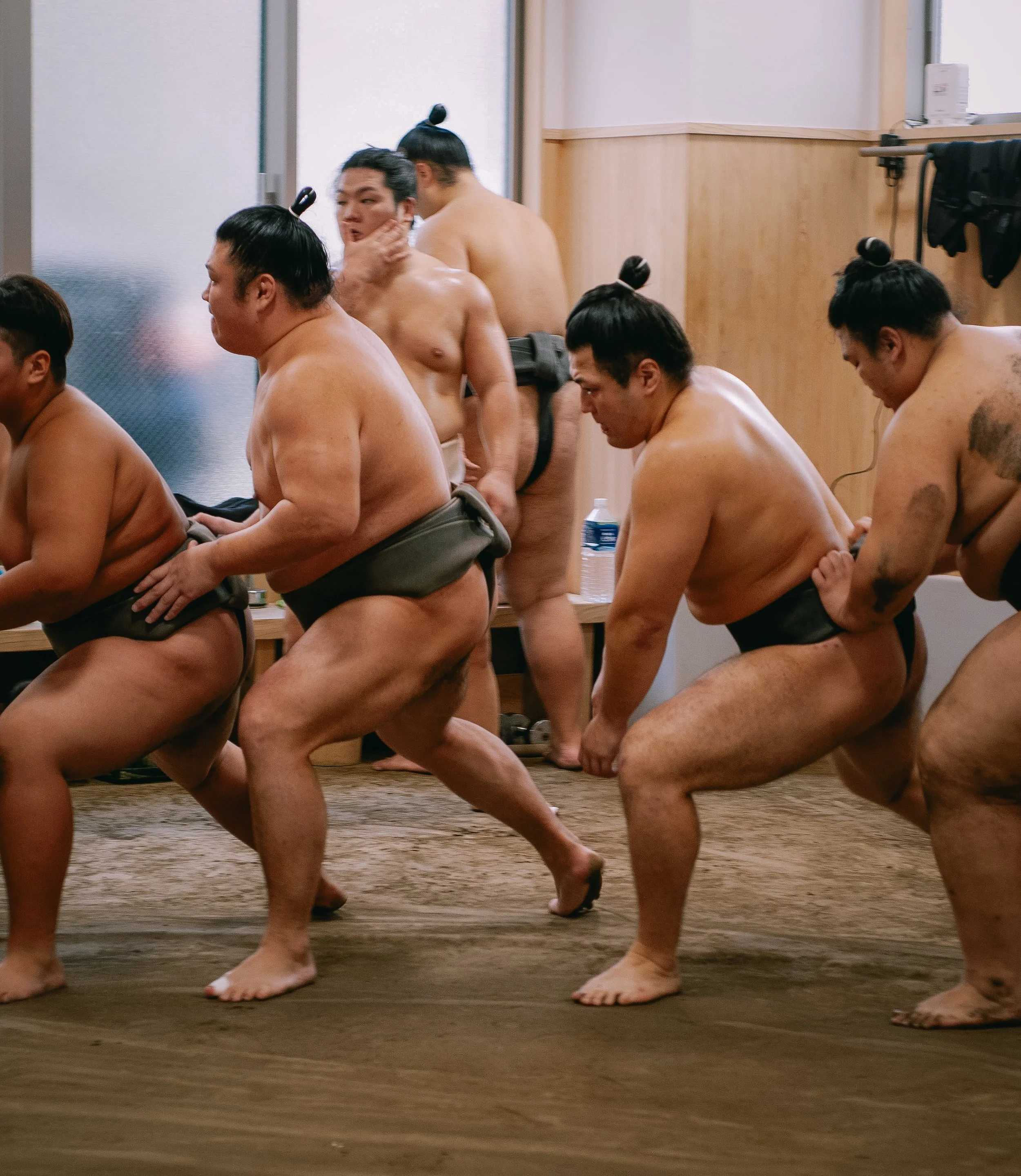 Sumo wrestlers in traditional mawashi engaging in a match inside a room with wooden walls, some sitting on a wooden bench and others standing in the background.