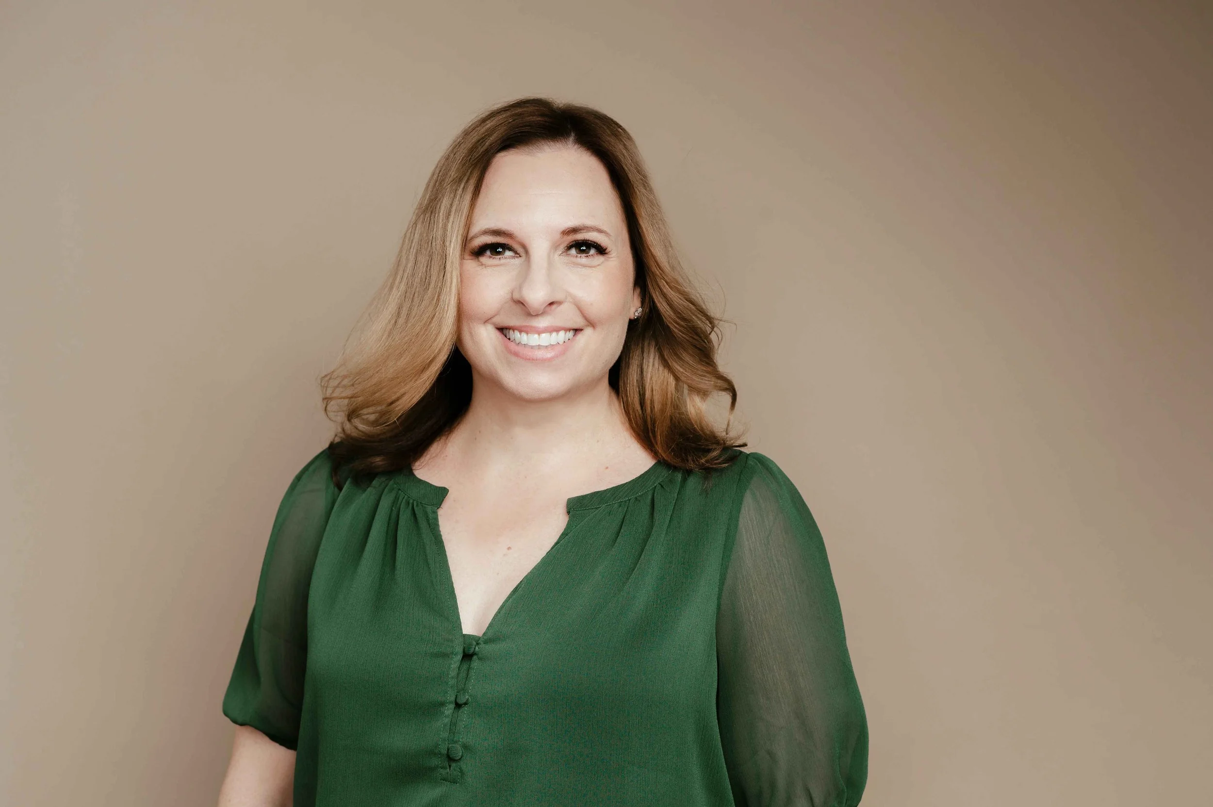 A woman with shoulder-length brown hair wearing a green blouse and smiling, standing against a beige background.