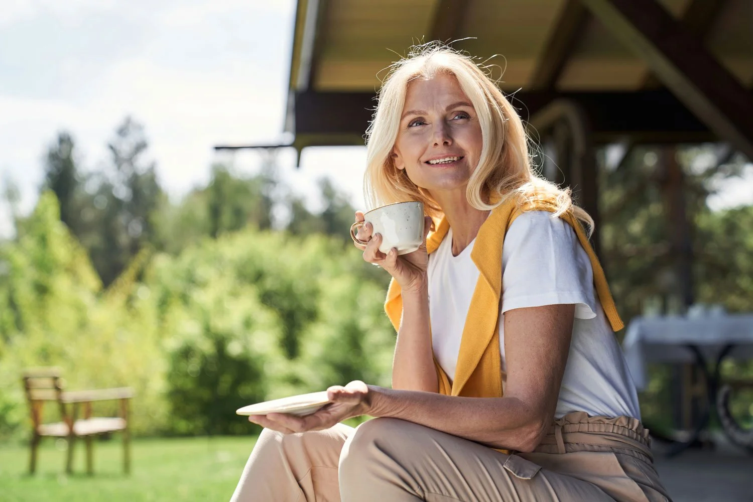A smiling woman with blonde hair sitting outdoors on a sunny day, holding a cup of coffee and a small plate, with a green garden and trees in the background.