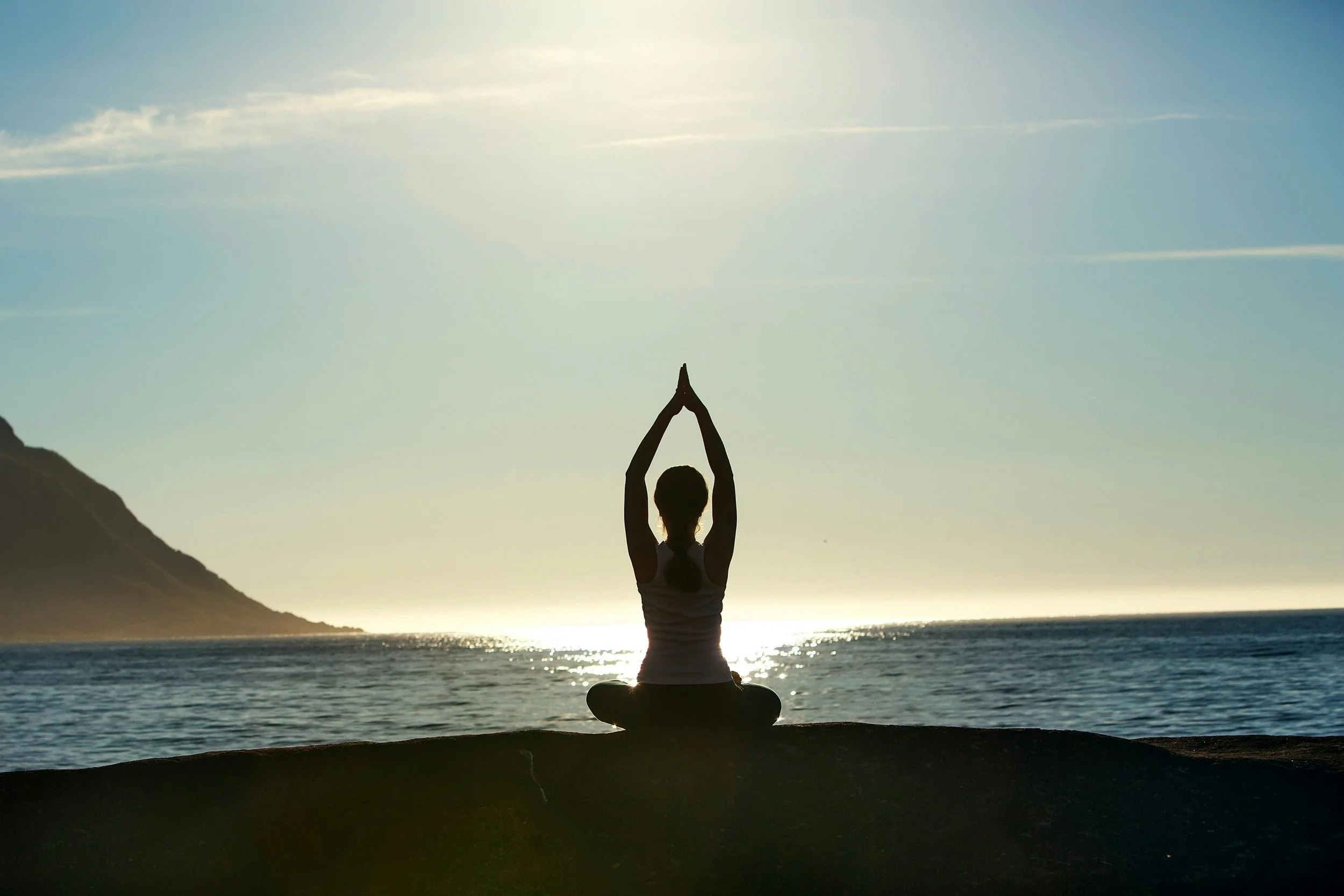 Person practicing yoga outdoors on a beach during sunset, sitting in a cross-legged position with hands in a prayer pose above the head.