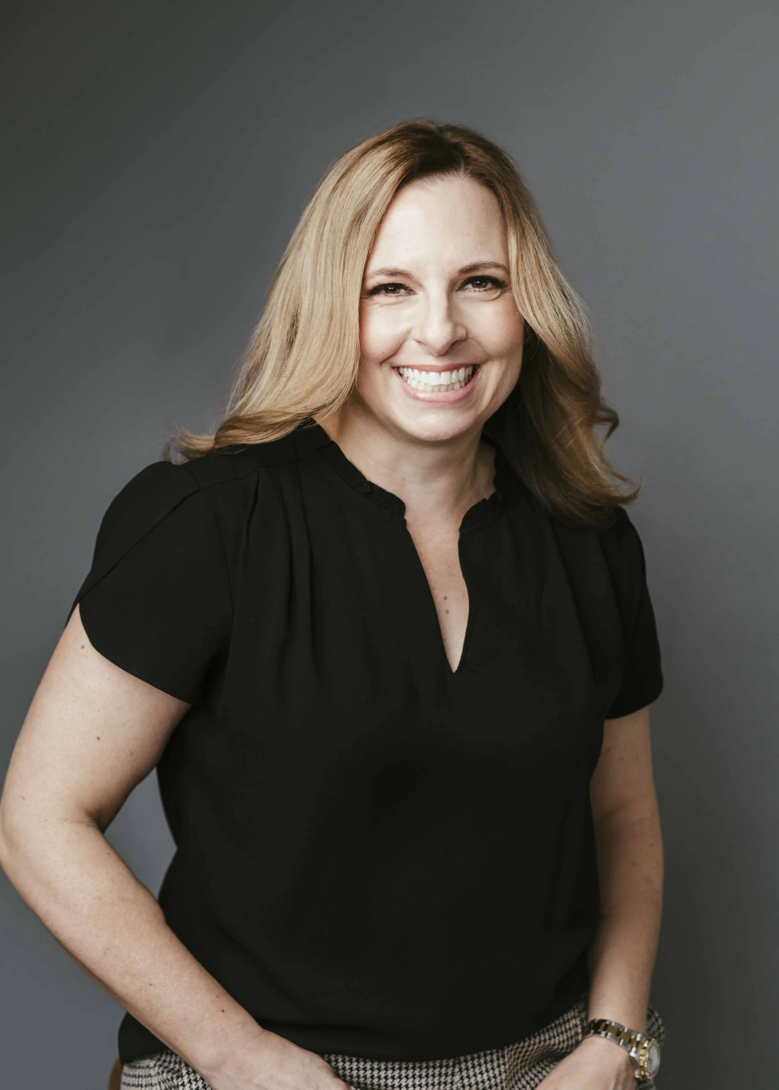A woman with wavy blonde hair, smiling, wearing a black top and a houndstooth checkered skirt, against a gray background.