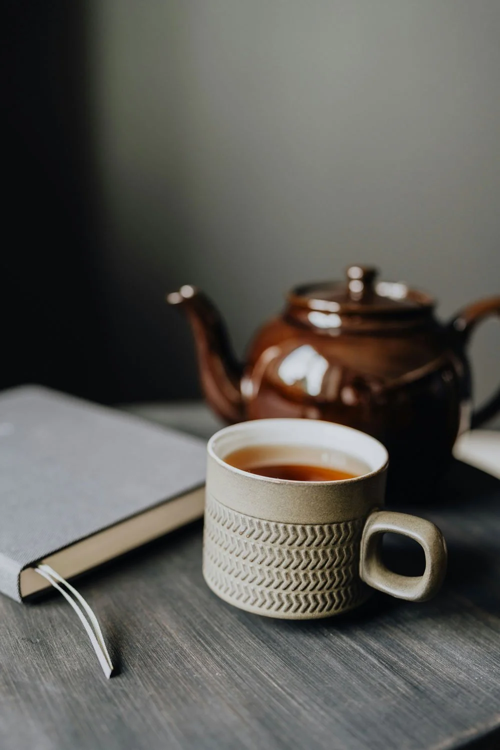 A beige ceramic mug filled with tea, placed on a gray wooden table next to a closed gray notebook, with a brown teapot in the background.