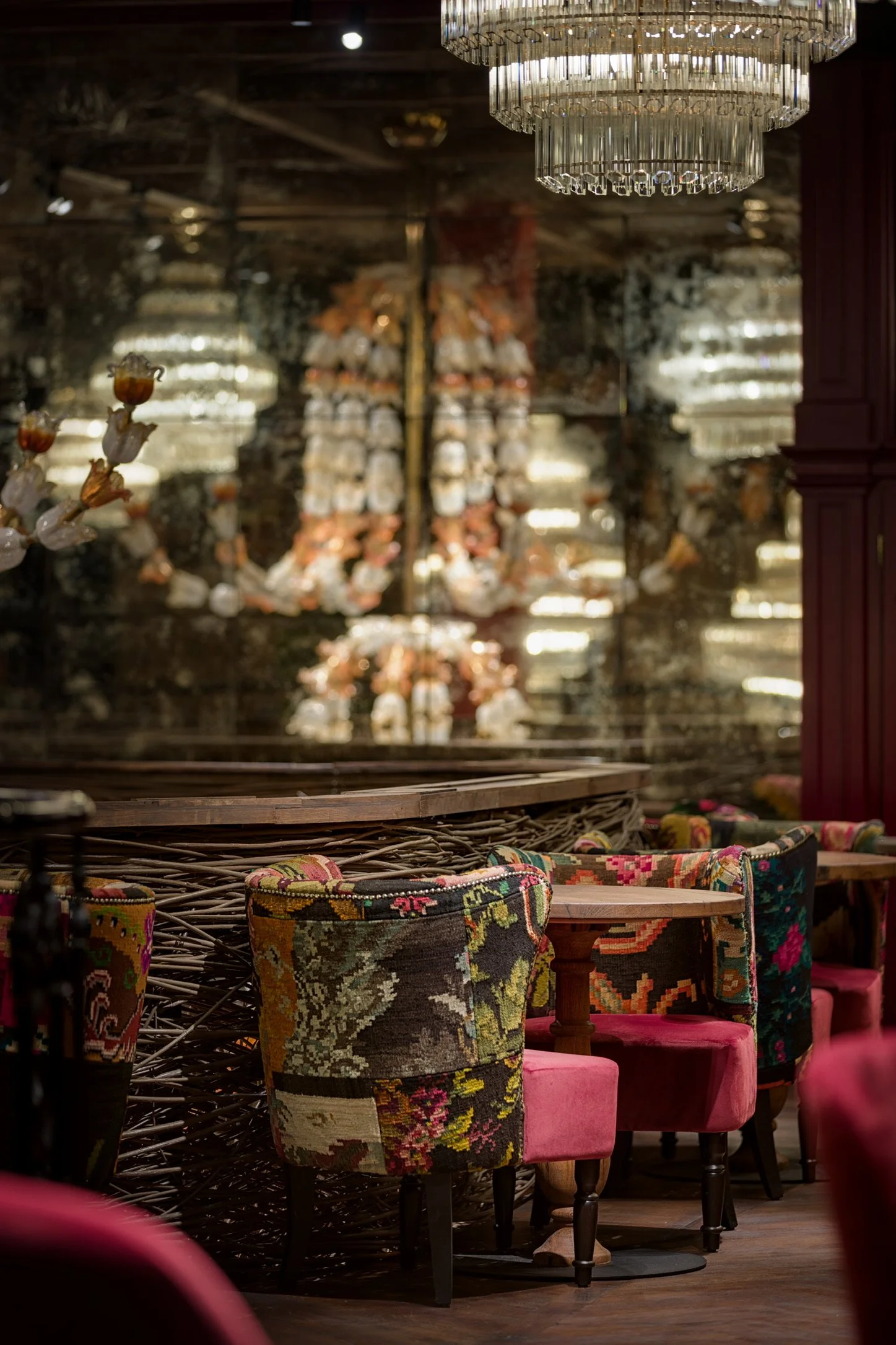 Chairs with floral patterned upholstery and pink velvet seats next to a wooden bar counter with a wicker front, reflected chandeliers, and a dark ornate mirror in the background.