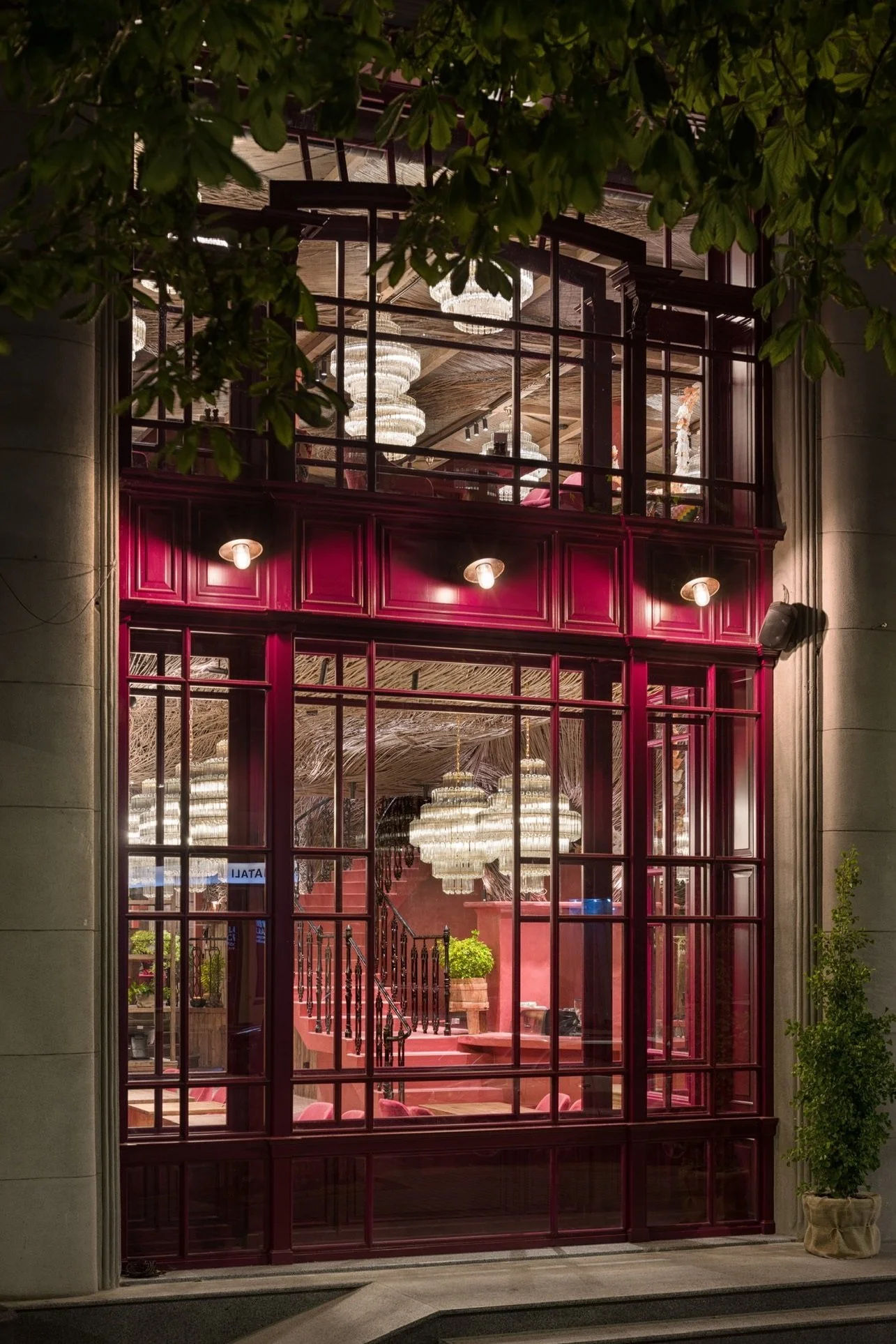 View through a large glass window of an interior space with chandeliers, a staircase with black railing, pink walls, and potted plants.