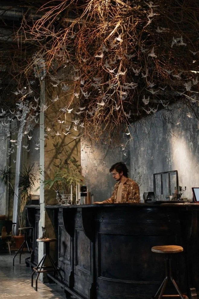 A man behind a dark wooden bar in a dimly lit cafe or restaurant, with a ceiling decorated with hanging white paper bats and dried branches, and potted plants along one wall.