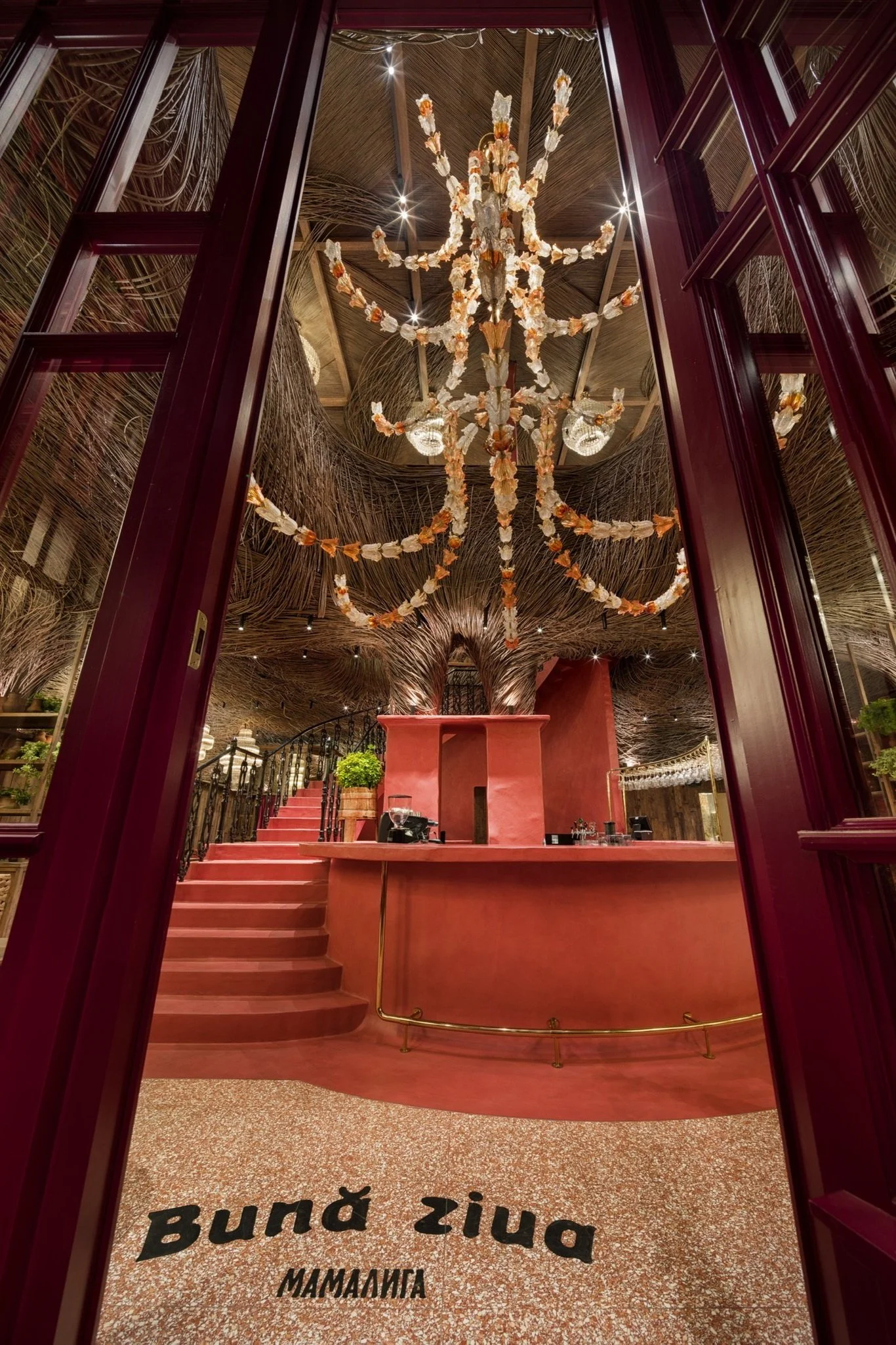 Interior view of a restaurant called Bunda Zina with pink and rust-colored decor, a grand chandelier decorated with seashells, a curved staircase, lush green plants, and intricate wood and straw accents.