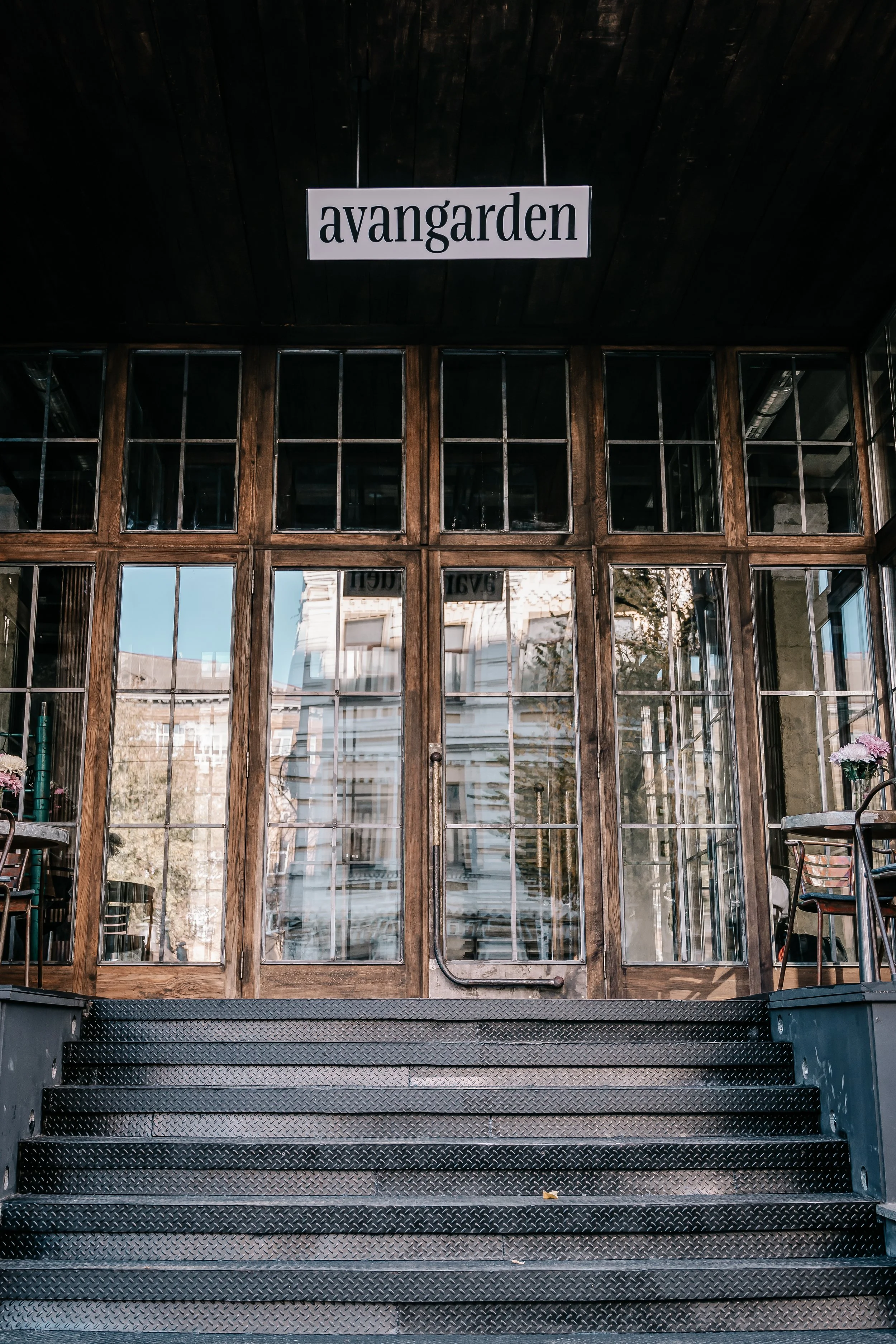 Front entrance of a building with large glass doors and windows, wooden frames, and a sign above that says 'avangarden'. There are metal stairs leading up to the entrance and some chairs and flowers visible inside.