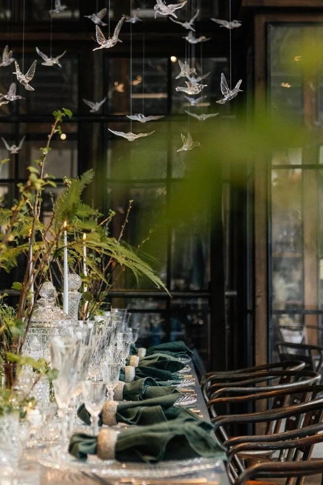 A dining table decorated with greenery, glassware, and green napkins with gold rings, set in a room with large windows and a hanging glass bird decoration.
