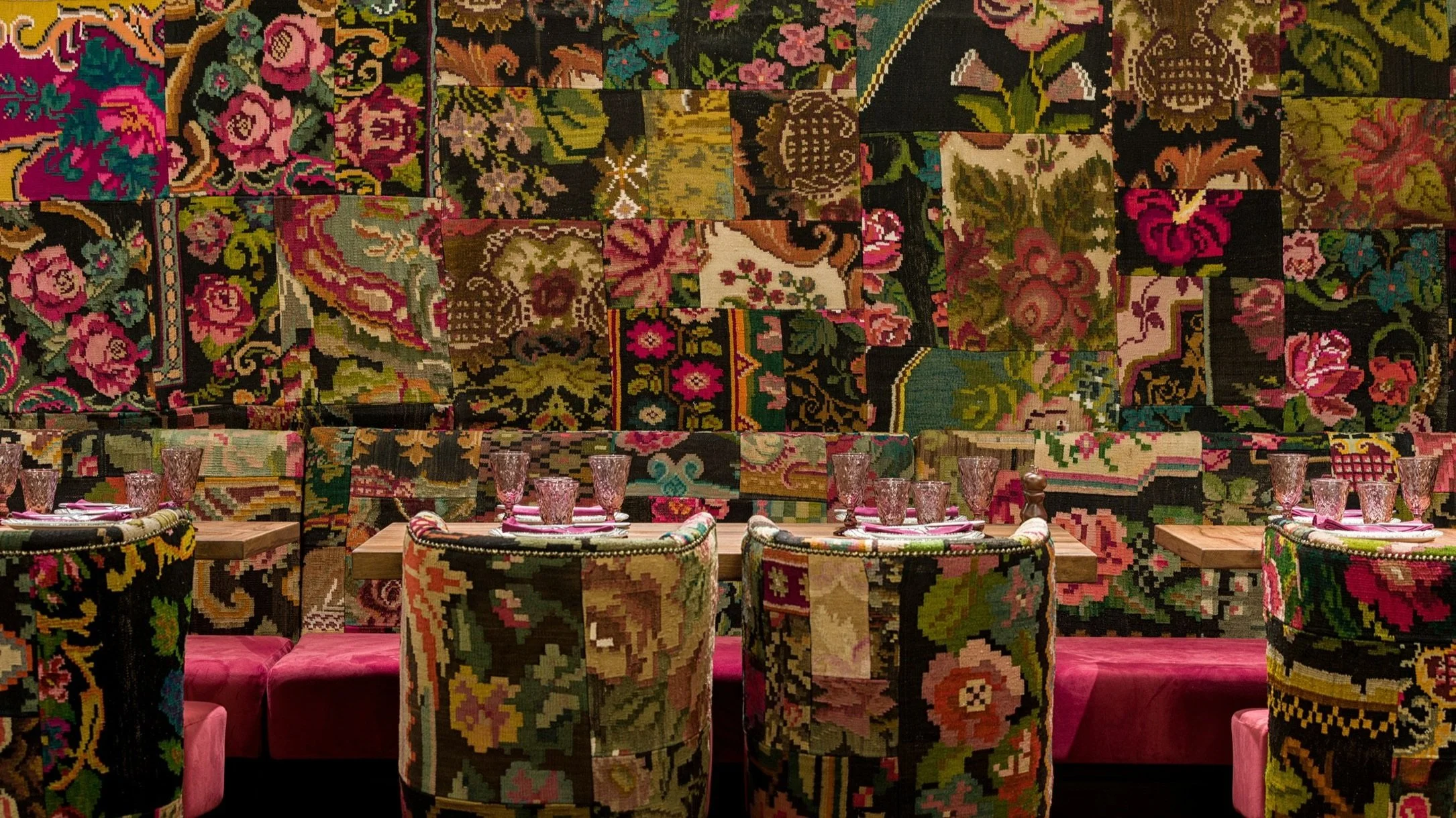 A dining area with tables and chairs, featuring a wall and chair upholstery with a patchwork of floral and decorative carpet patterns, creating a vibrant and eclectic ambiance.