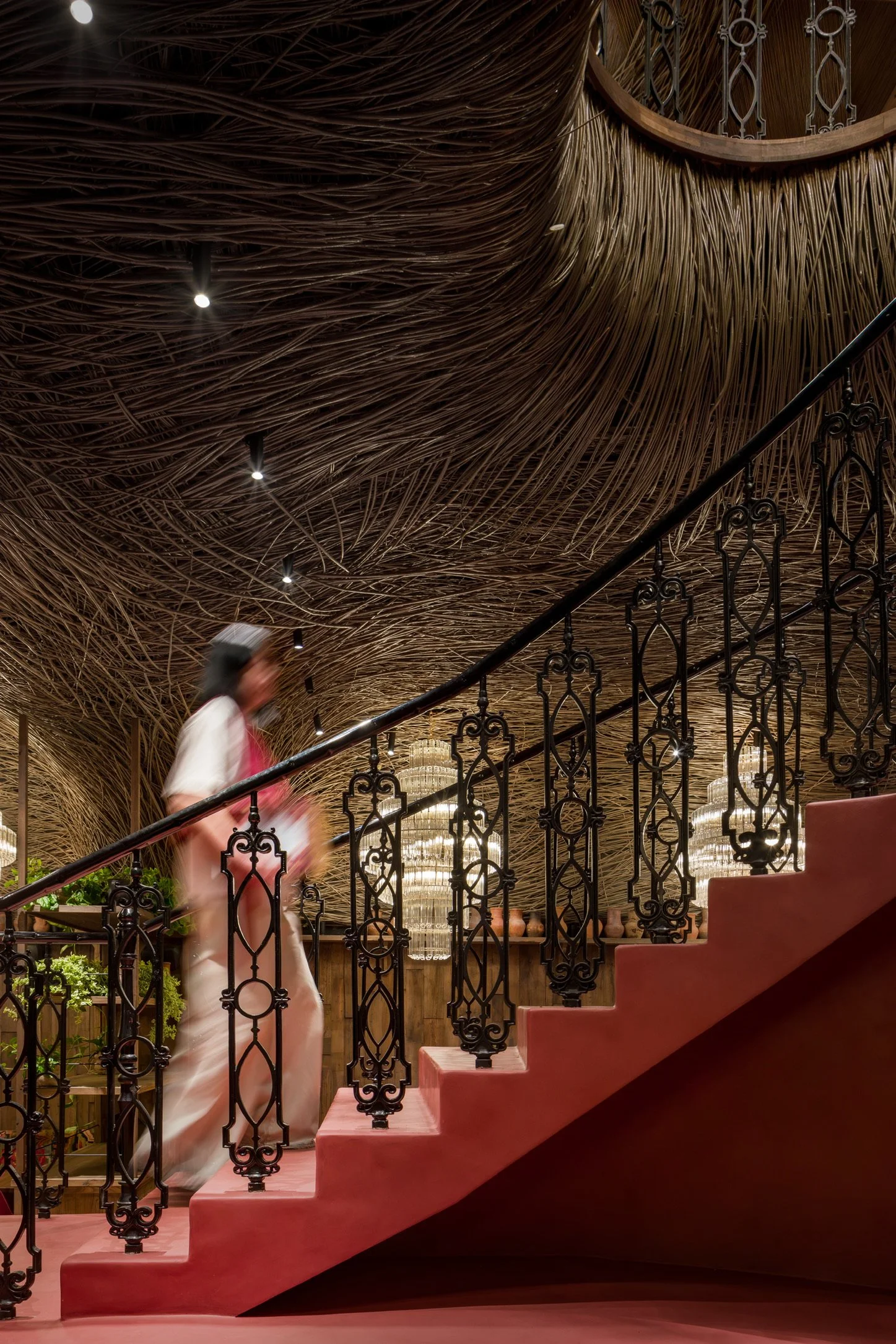 Interior of a building with a woven reed ceiling, ornate black metal staircase railing, pink stairs, a blurred person walking up, and chandeliers in the background.