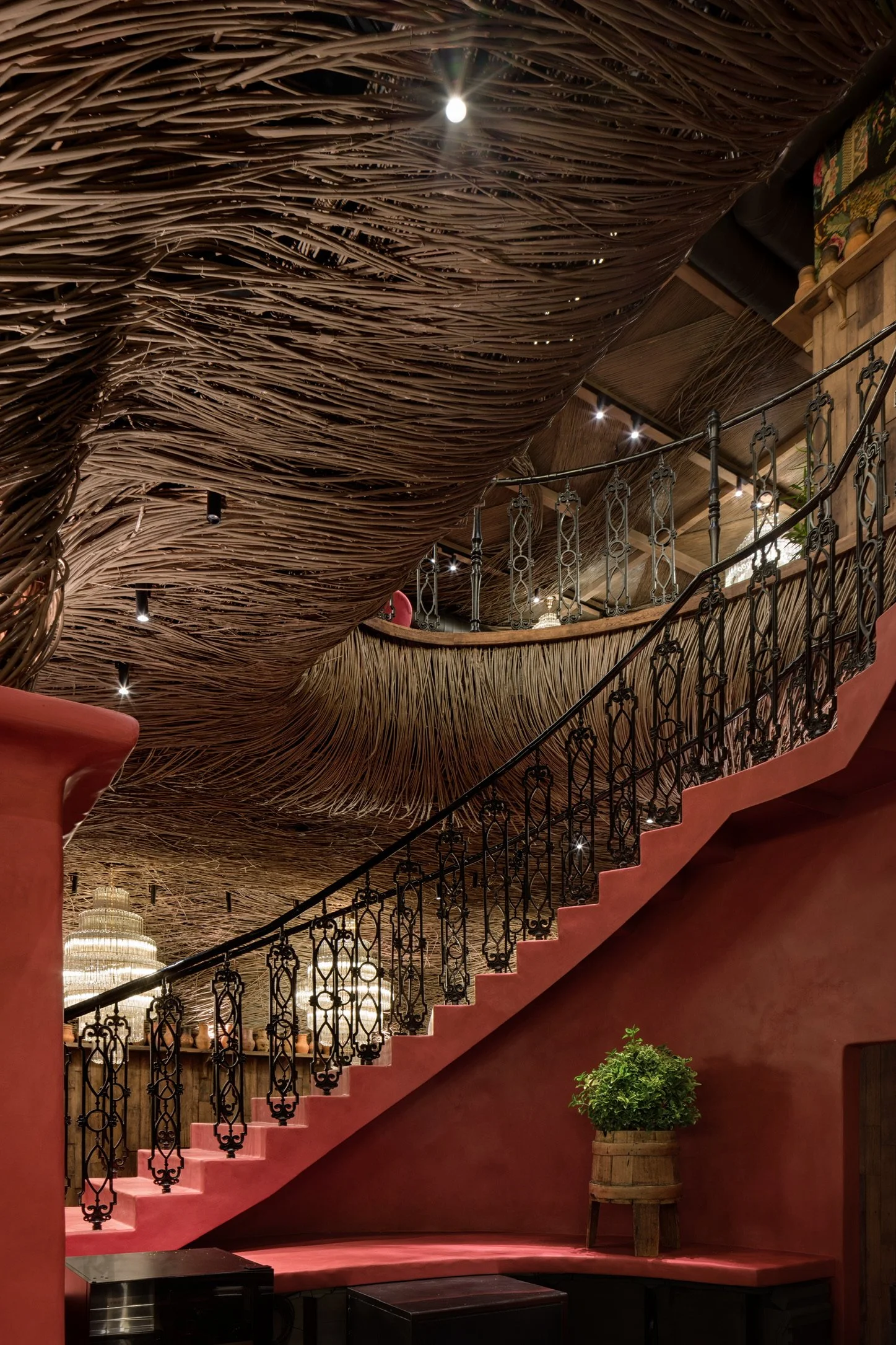 Interior view of a restaurant or café with a unique ceiling made of intertwined twigs or reeds, a staircase with ornate black metal railing, red stairway, and a pink wall with a small green plant in a wooden barrel.