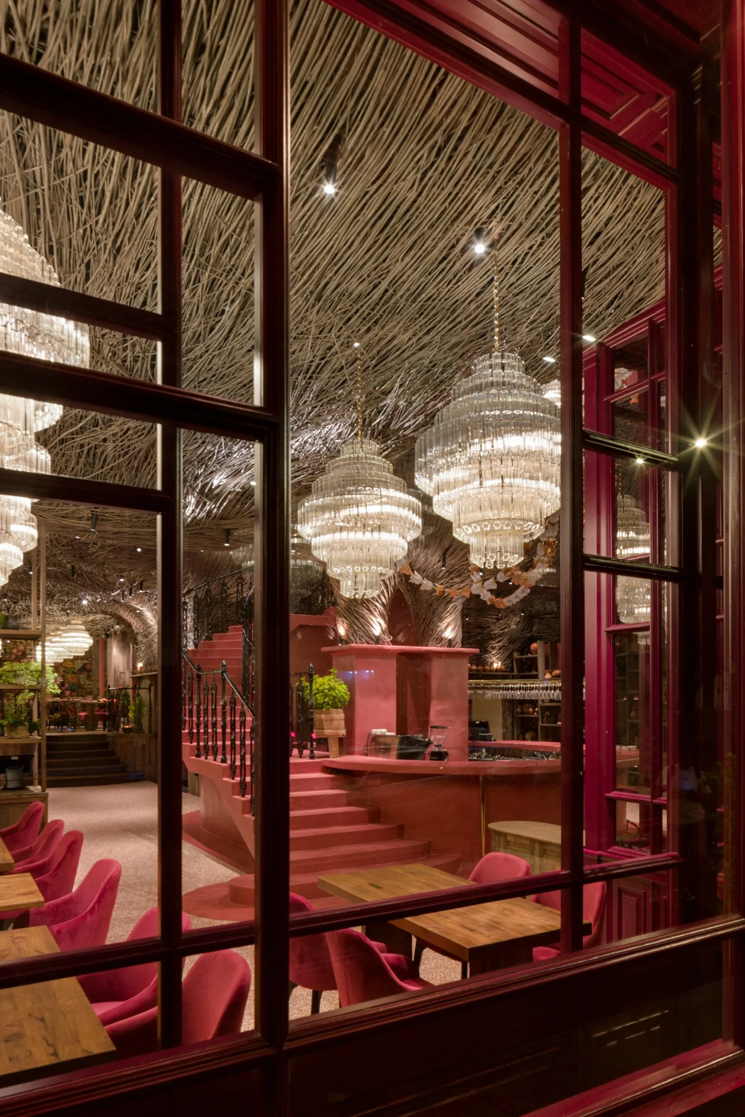 Interior of an elegant restaurant or lounge with chandeliers, pink furniture, and a thatched ceiling seen through windowpanes.