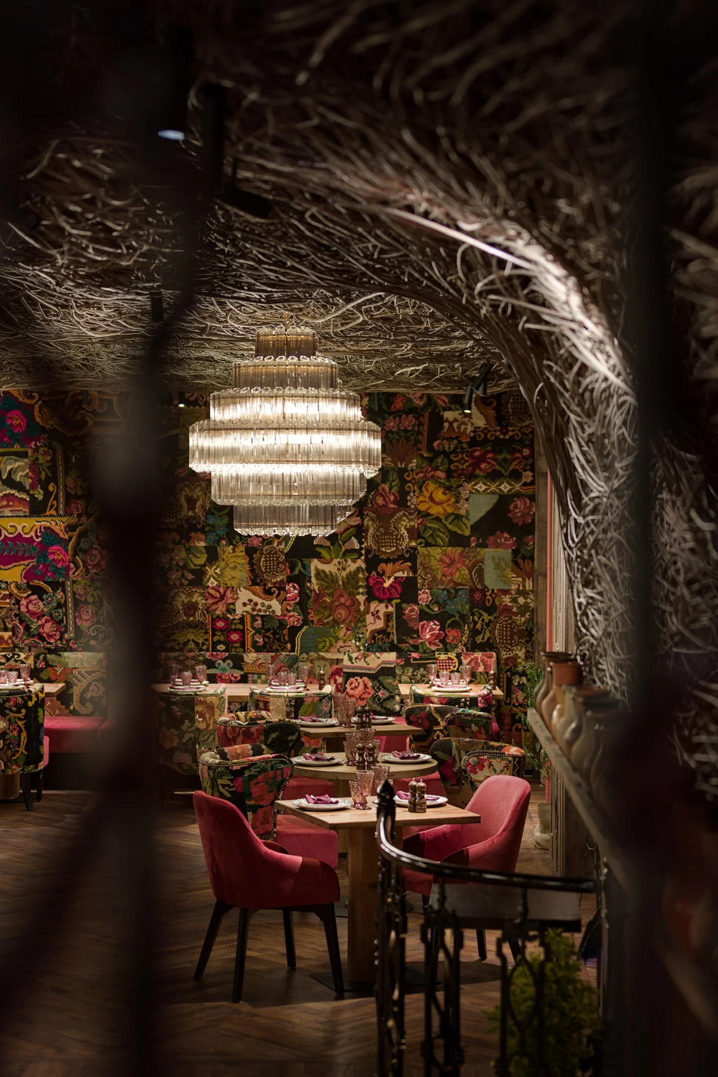 Elegant restaurant interior with ornate woven ceiling, colorful floral wallpaper, round tables set with glasses, plates, and napkins, surrounded by pink and patterned chairs, illuminated by a large chandelier.