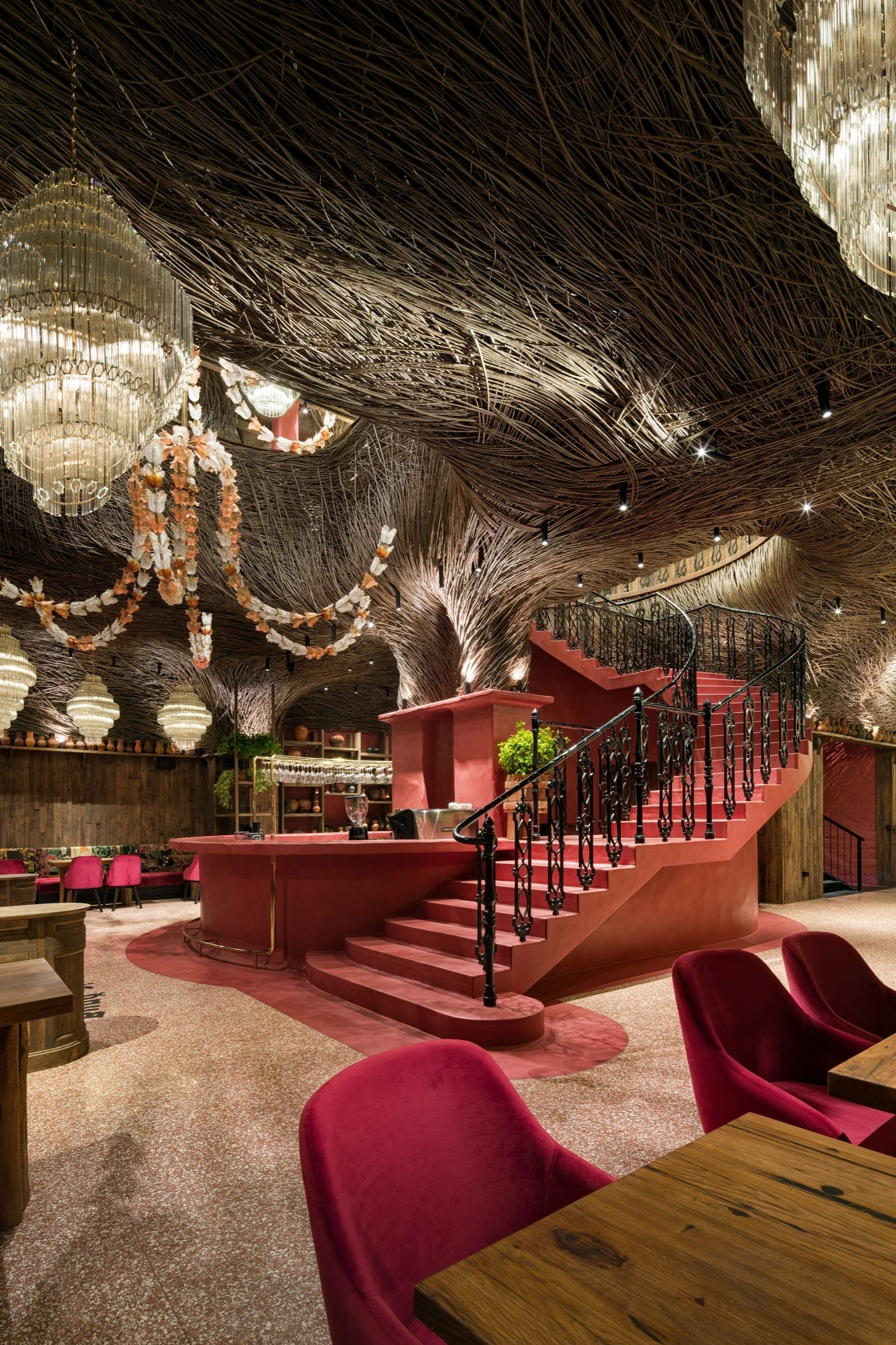 An elegant restaurant interior decorated with chandeliers, pink and wood accents, a central bar area with pink stairs, and a ceiling designed to resemble intertwined branches.