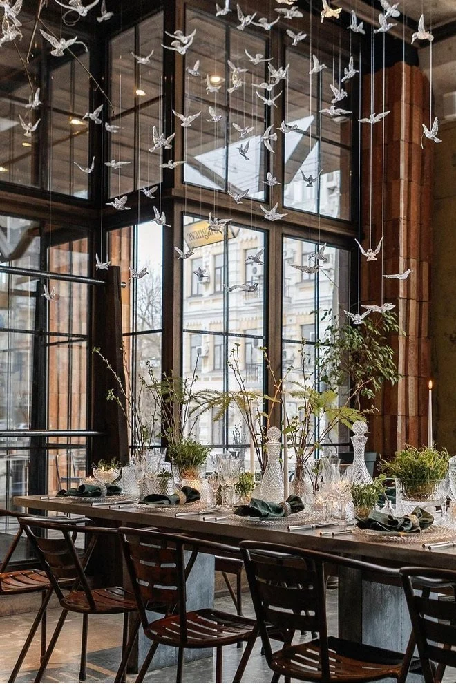 Indoor dining table decorated with glassware and floral centerpieces, surrounded by wooden chairs, with large windows and hanging paper bird decorations overhead.
