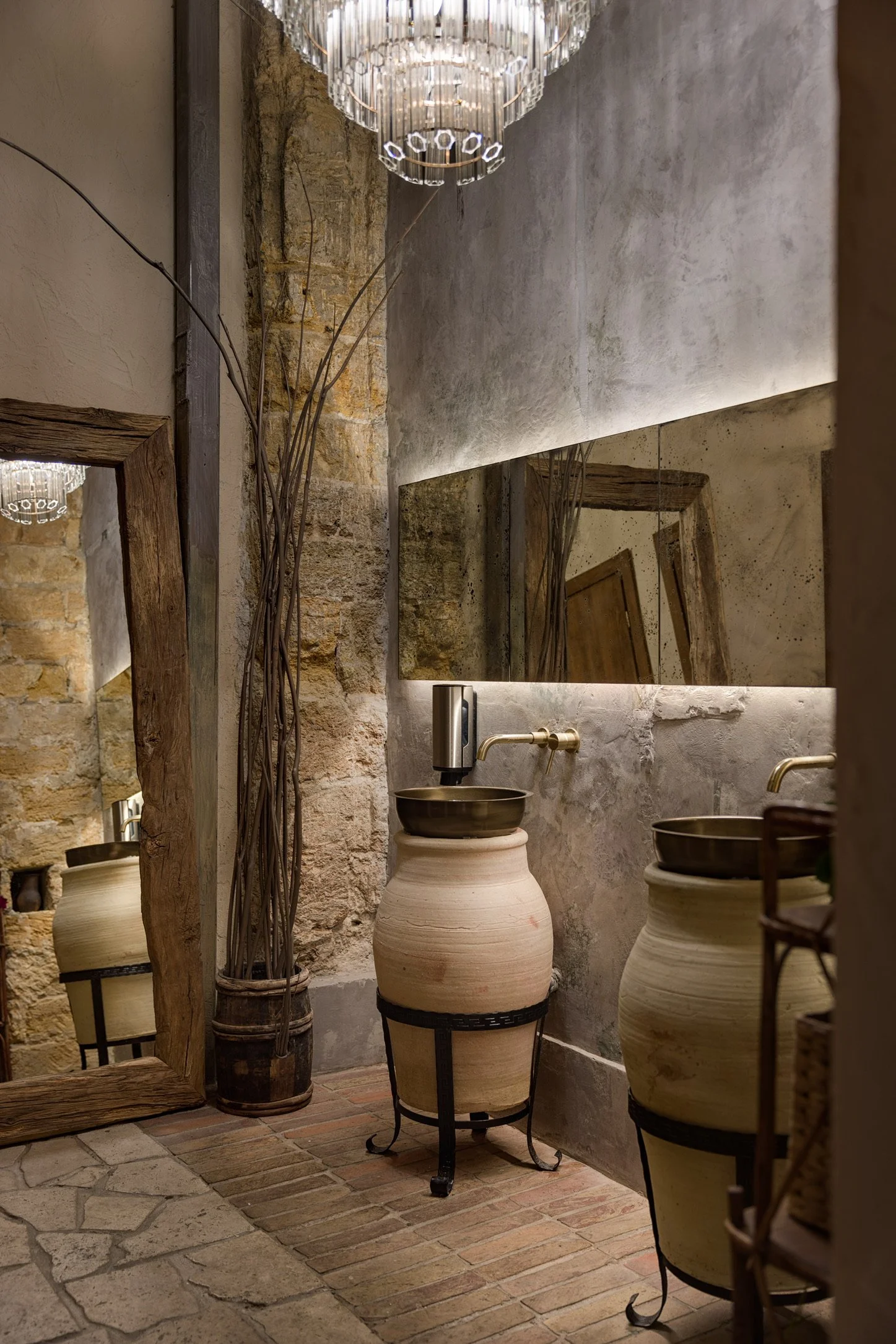 A rustic bathroom with two ceramic washbasins atop black metal stands, large mirrors, a chandelier, brick and stone walls, and dried branches in a vase.