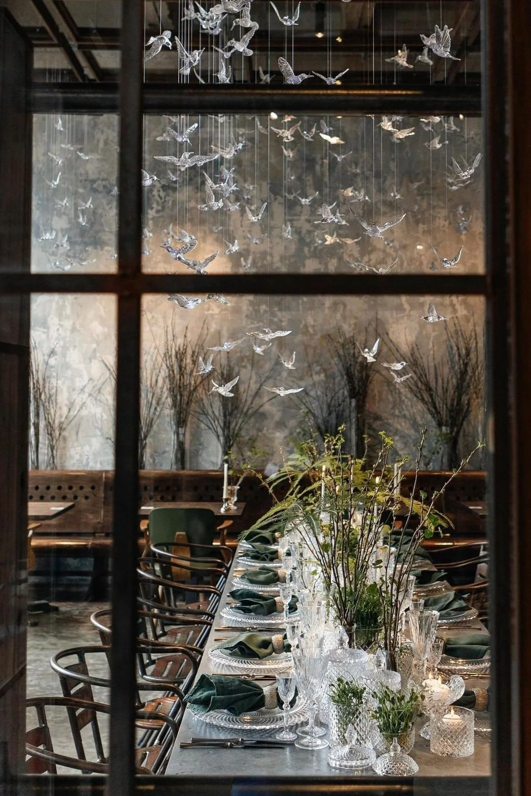Elegant dining room viewed through a window, featuring a long table set with crystal glassware, dark green napkins, and floral centerpieces with branches and greenery. The background has a textured wall with artwork or lighting patterns and a suspended installation of numerous glass or crystal birds hanging from the ceiling.