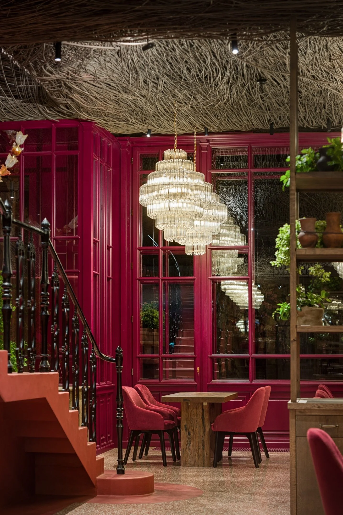 Elegant restaurant interior with a chandelier, pink walls, a wooden table, pink chairs, and decorative plants.