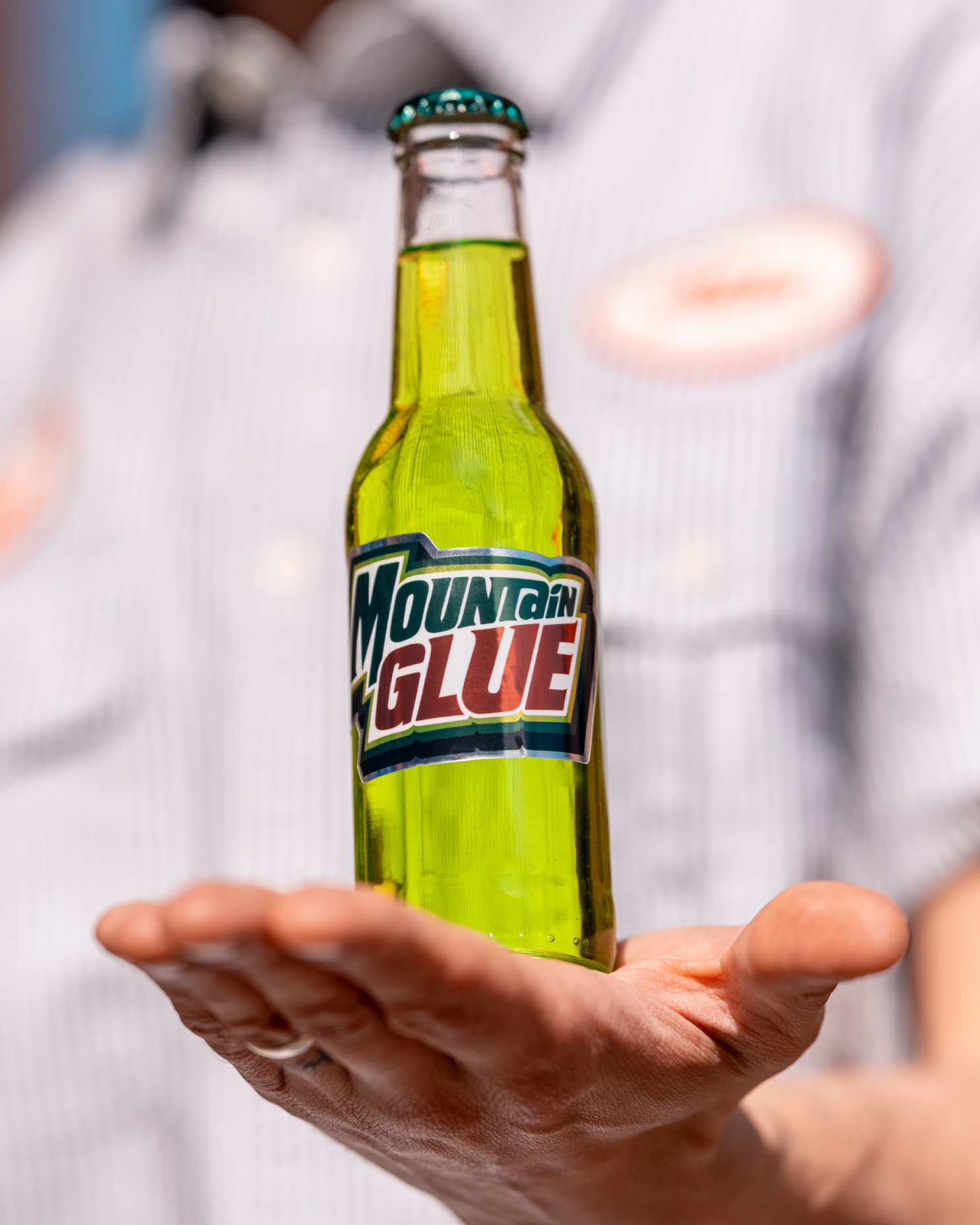 A person holding a clear glass bottle of Mountain Glue with a greenish-yellow liquid inside.