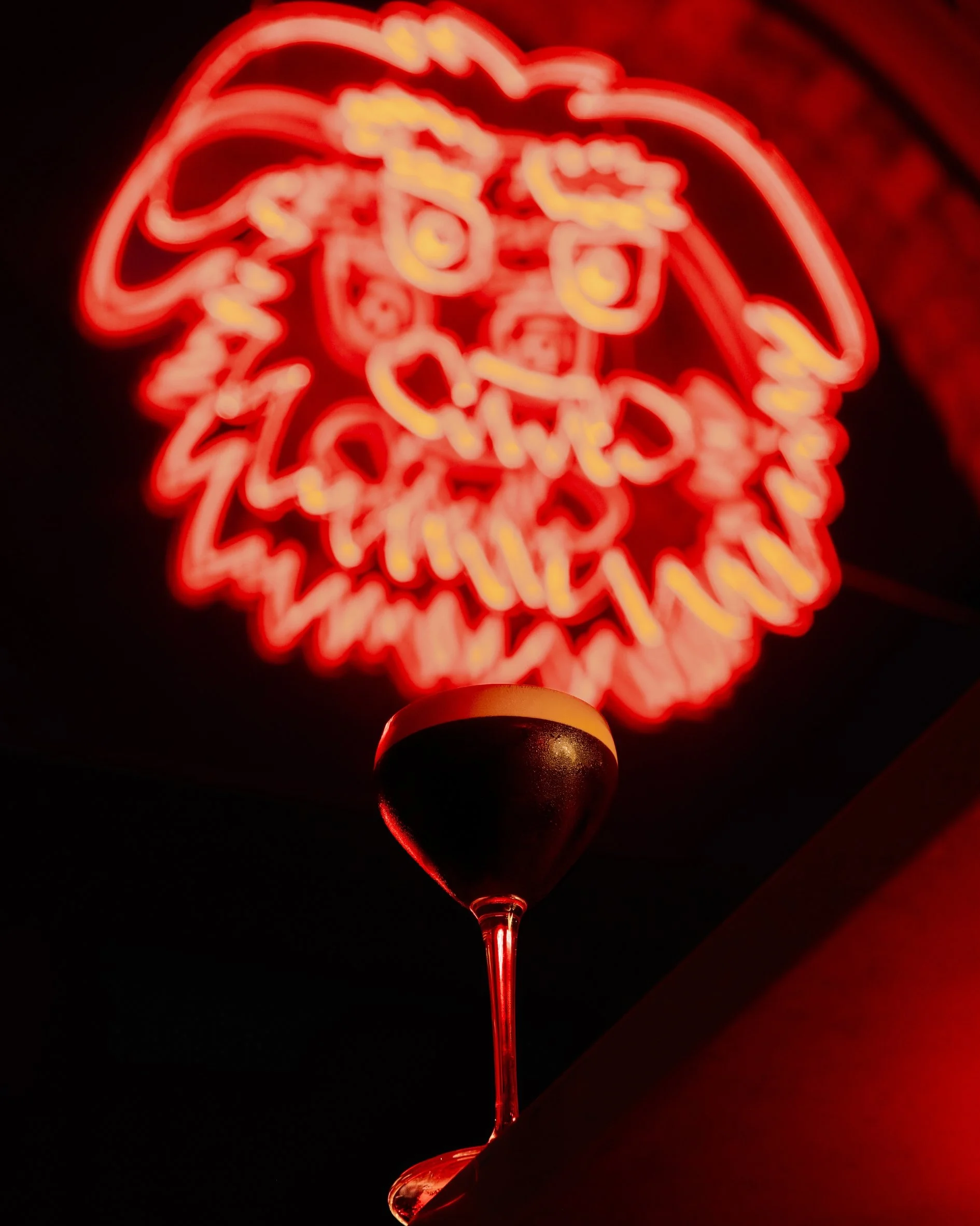 Neon sign with a red painted lady graphic and the words 'Lola's Drinkery' in yellow, mounted on a dark wall.