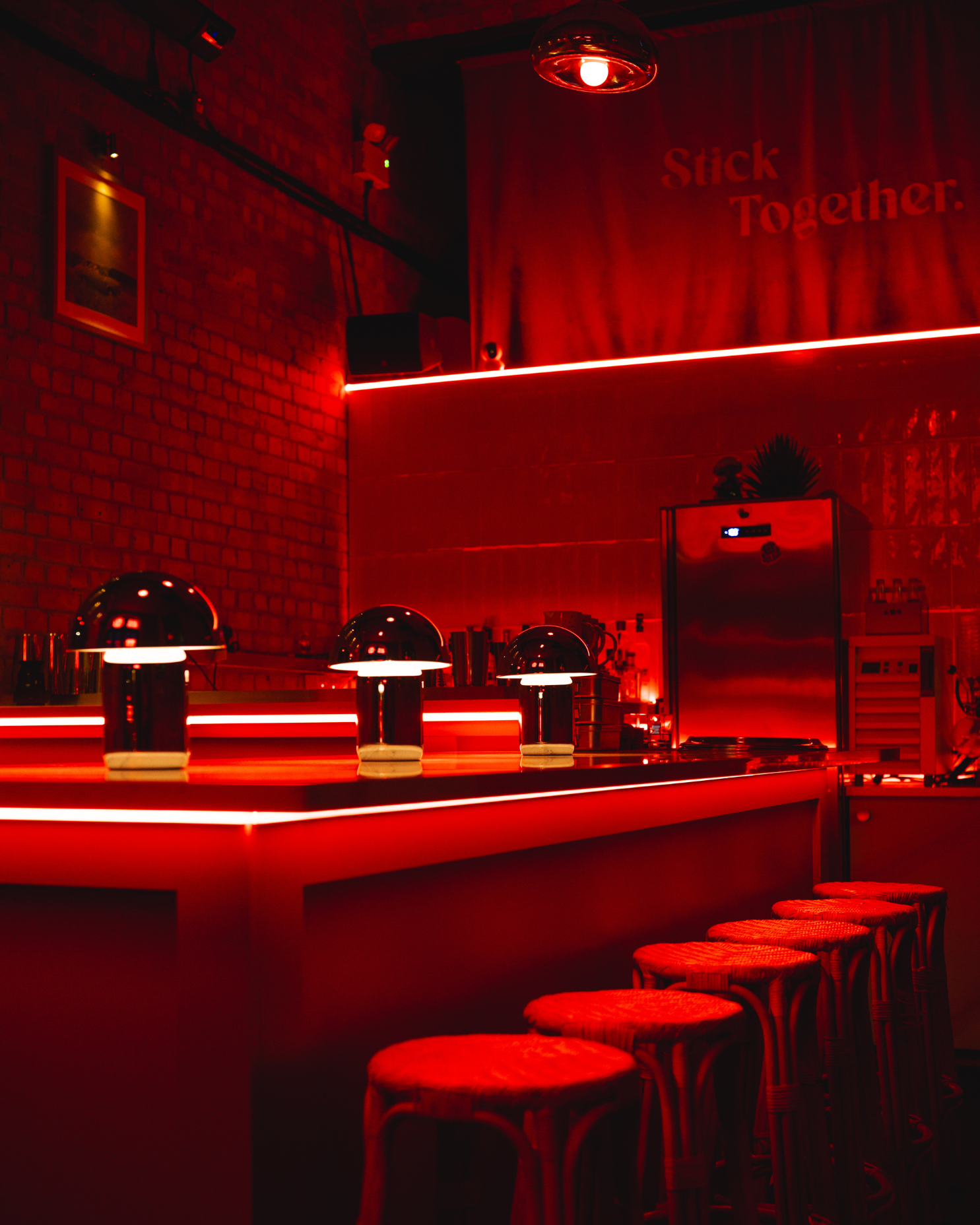 Dimly lit bar or lounge with red neon lighting, a row of stools along the counter, and a brick wall with framed art.