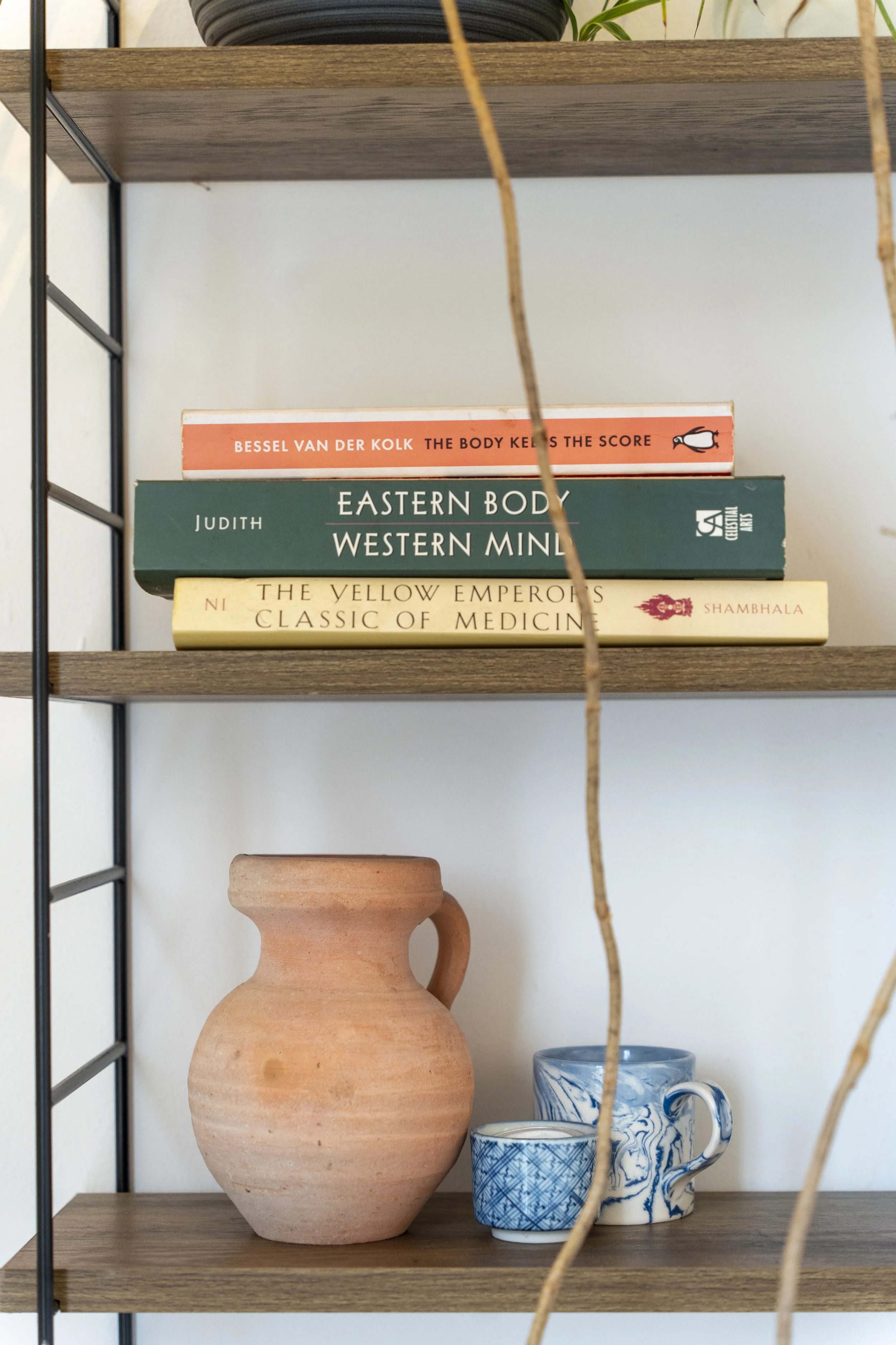 A wooden bookshelf with three books stacked on top. Below, there is a small terracotta pitcher and two blue and white ceramic cups.