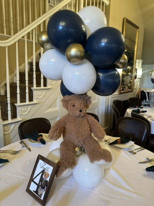 Table centerpiece with a teddy bear, white, navy, and gold balloons, and a framed photo or card, in a decorated dining room.