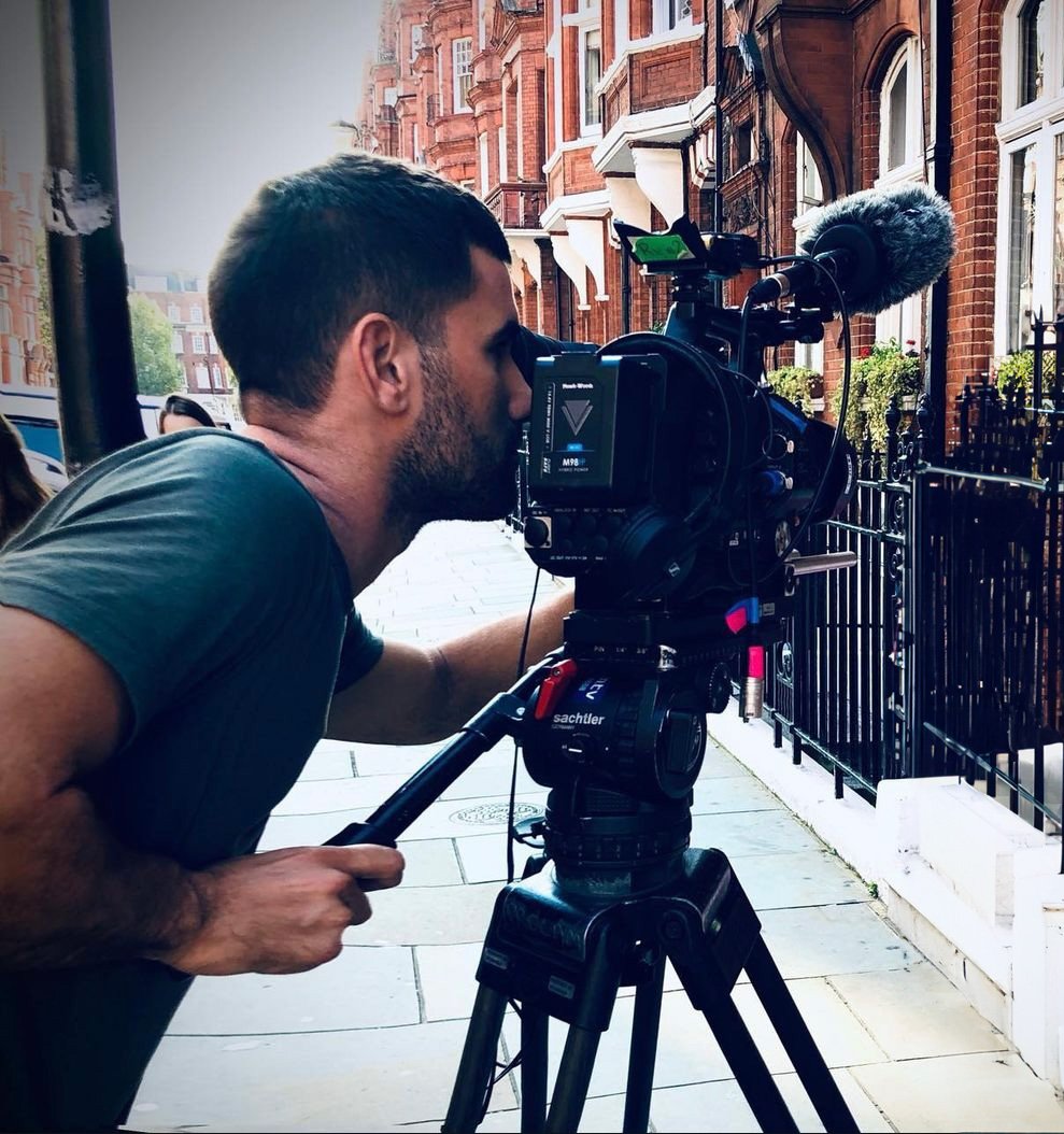 A man operating a professional video camera on a tripod on a city sidewalk with red-brick buildings and black iron fences in the background.