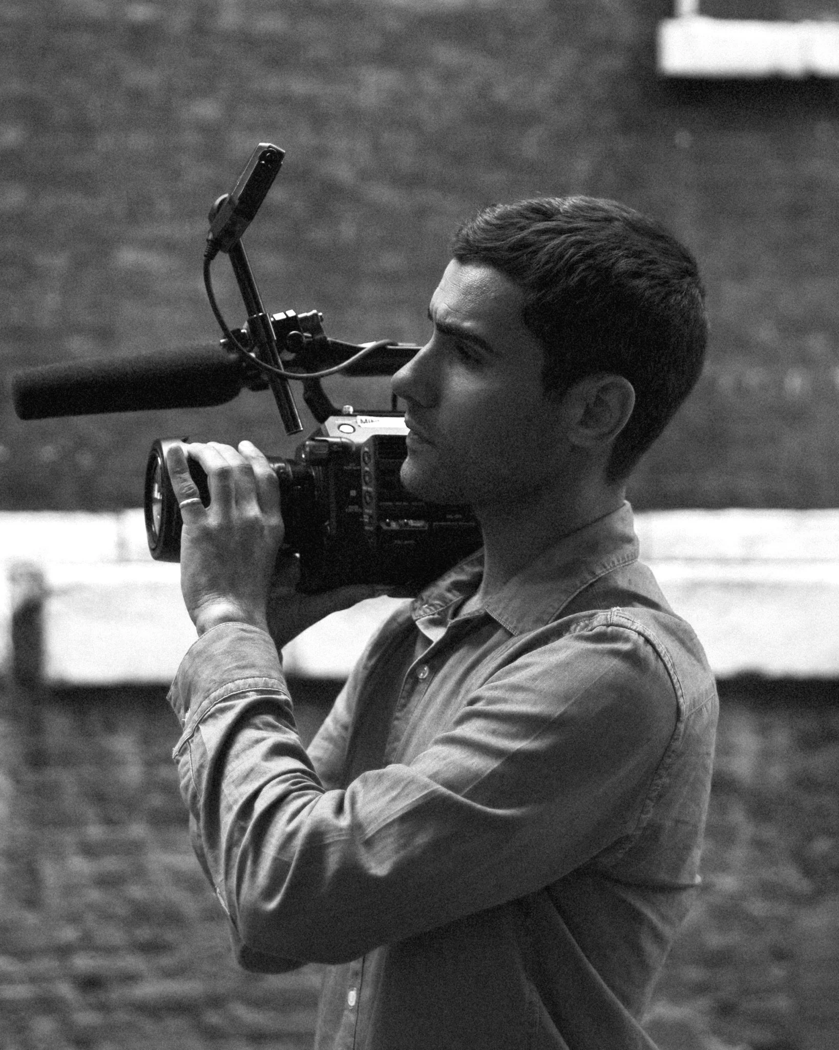 A man holds a professional video camera on his shoulder, recording or filming outdoors.