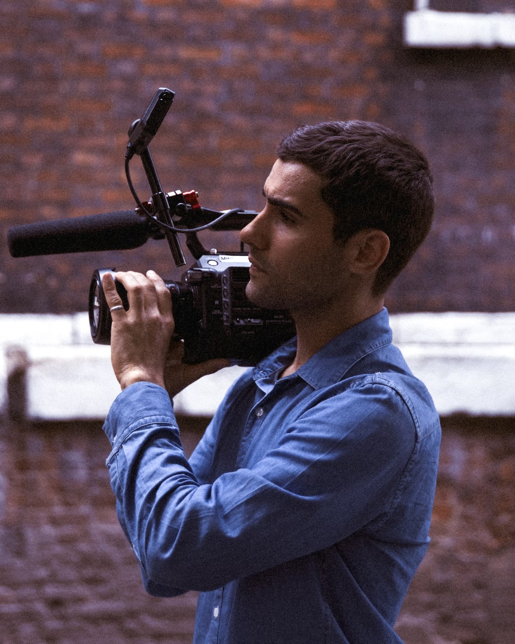 A man in a blue shirt is holding a professional video camera, filming or taking a photo against a brick wall background.