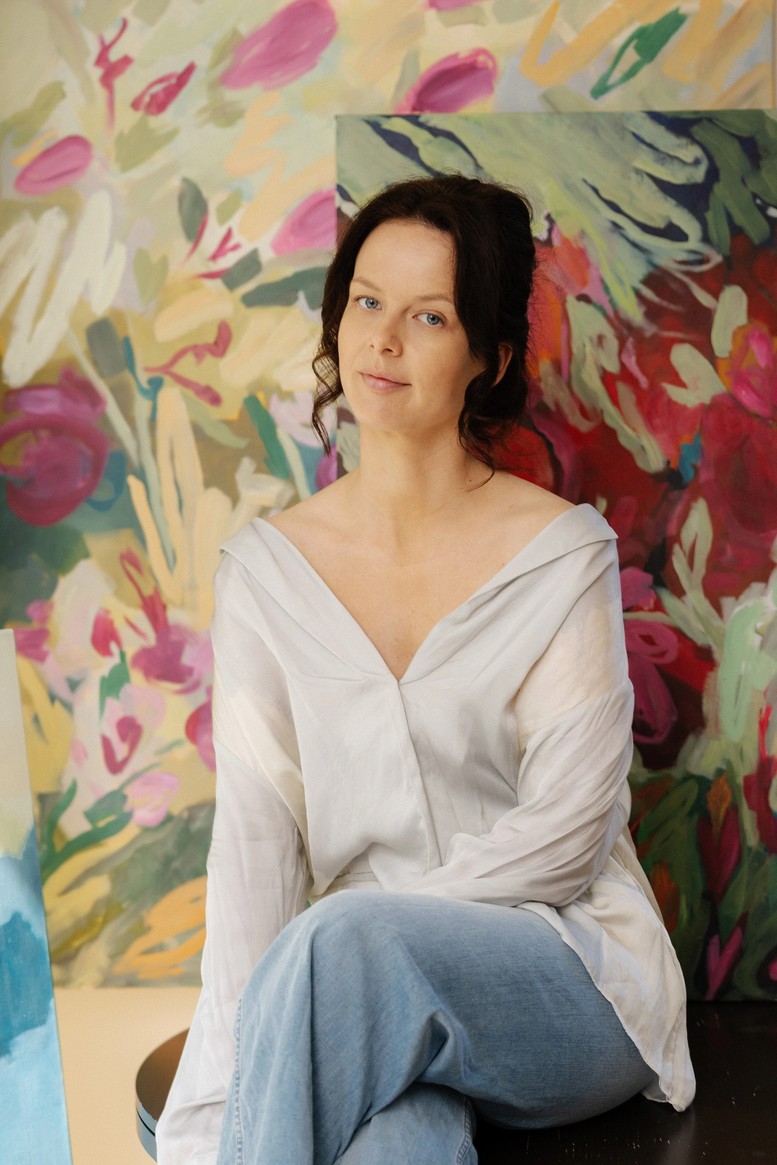 Contemporary visual artist and painter Kylie Hutton Herman sits with two abstract large original paintings. Features a woman artist with blue eyes, dark curly hair,  light shirt and jeans, in artist art studio based in Canada and The Bahamas.