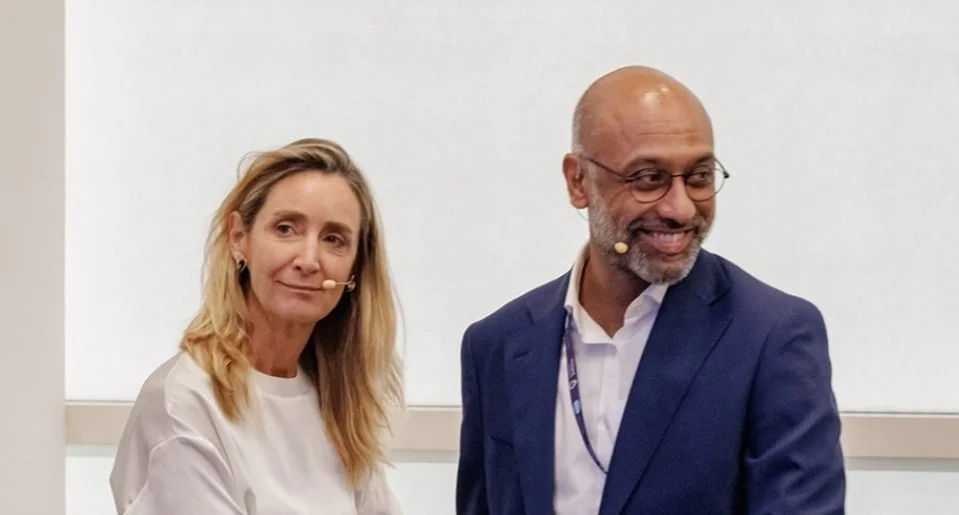 A woman and a man smiling, both wearing microphones during a presentation or conference.
