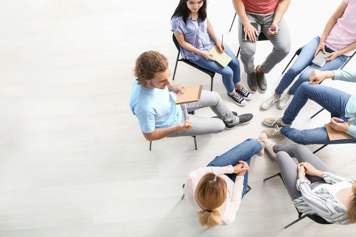 A group of people sitting in a circle during a therapy or support group meeting, with one person speaking while others listen and take notes.