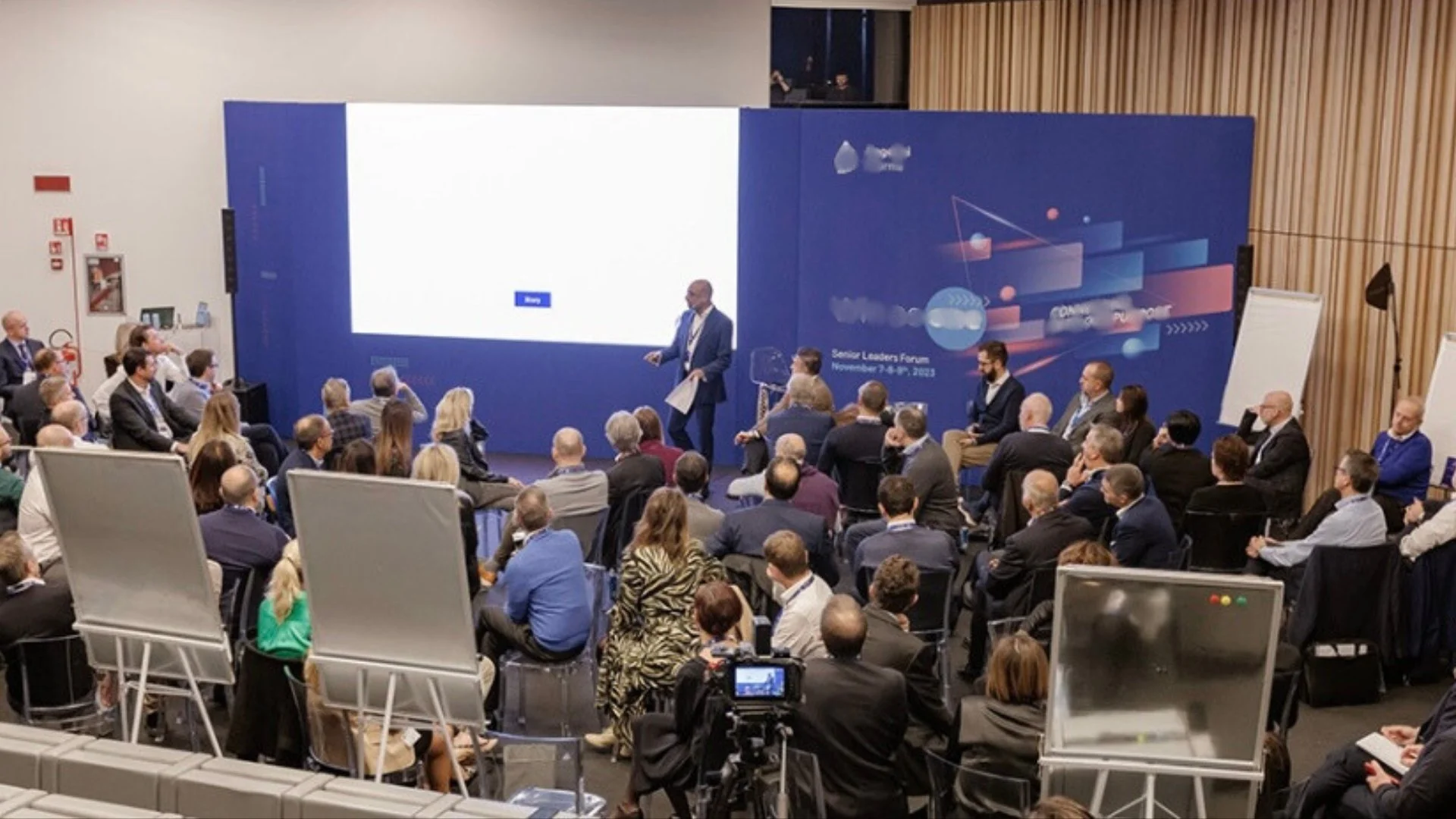 A large conference room filled with a diverse audience seated and listening to a speaker standing at a podium at the front of the room. There are big screens behind the speaker and whiteboards on the sides. The event appears to be a professional forum or seminar.
