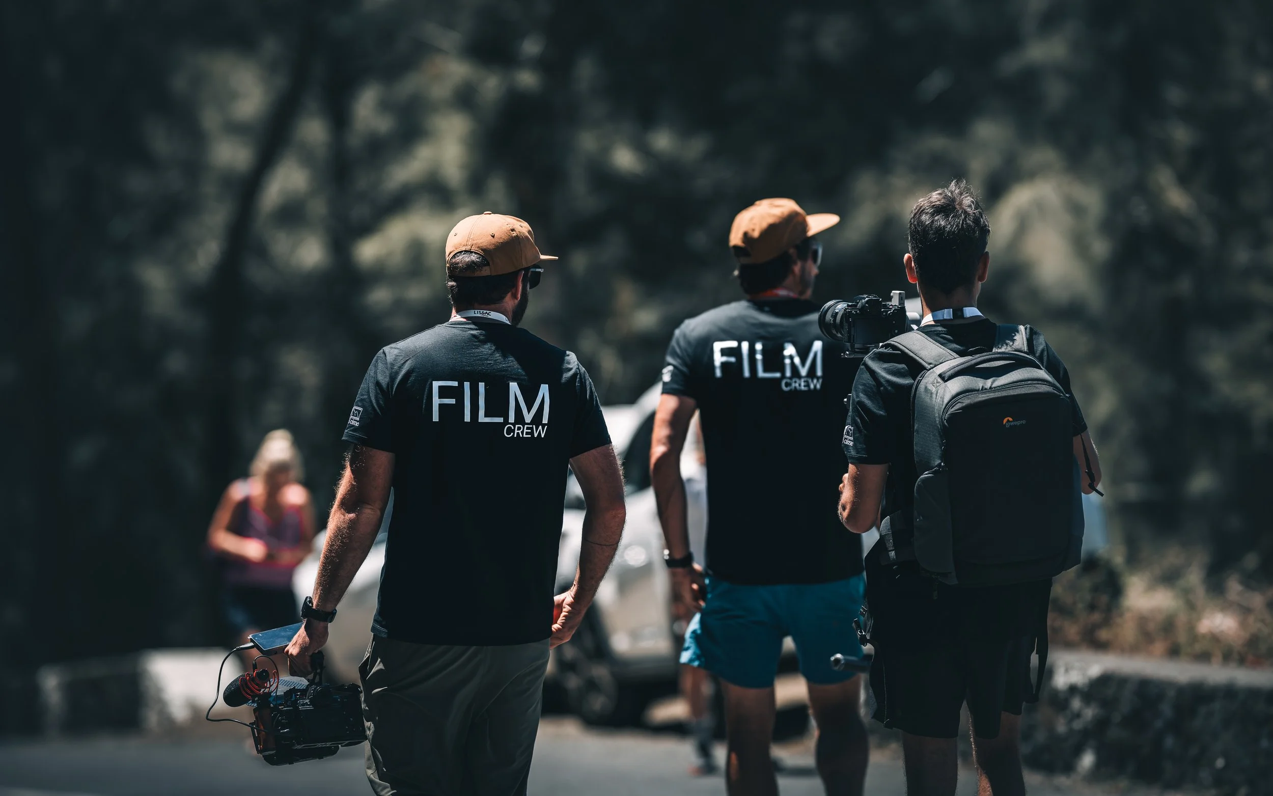 Filmmakers wearing black t-shirts with text "film crew" walking holding videocamera gear
