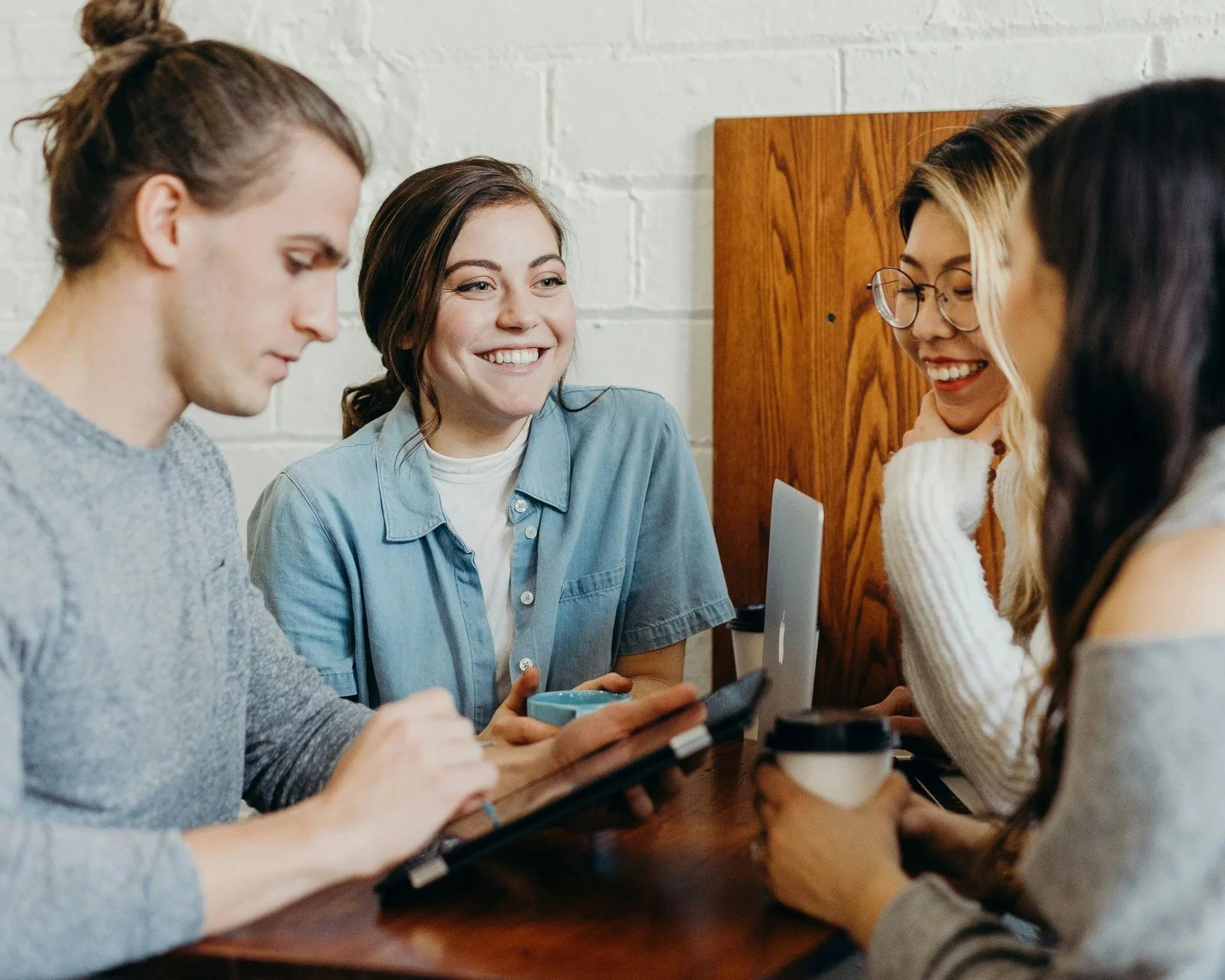 A group of five young women sitting around a wooden table with a white brick wall in the background. They are engaged in a conversation, smiling, and looking at each other. One woman in the middle is holding a smartphone. There are cups on the table, and one woman has her hand near her chin while smiling.