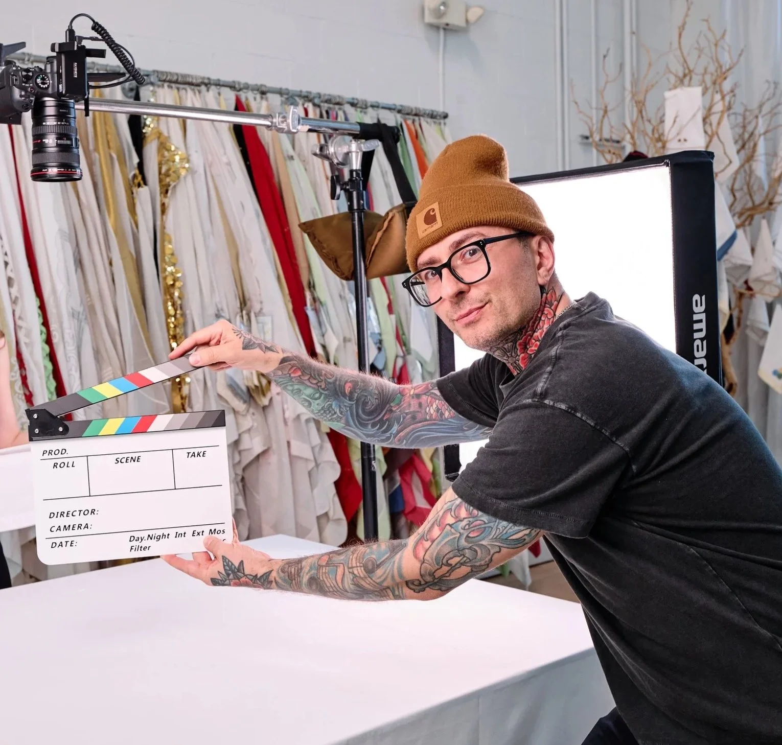 A man with tattoos wearing glasses, a brown beanie, and a black t-shirt is holding a film clapperboard in a clothing store. There is a professional light behind him, and racks of colorful clothing behind, with some decorative branches on the wall.