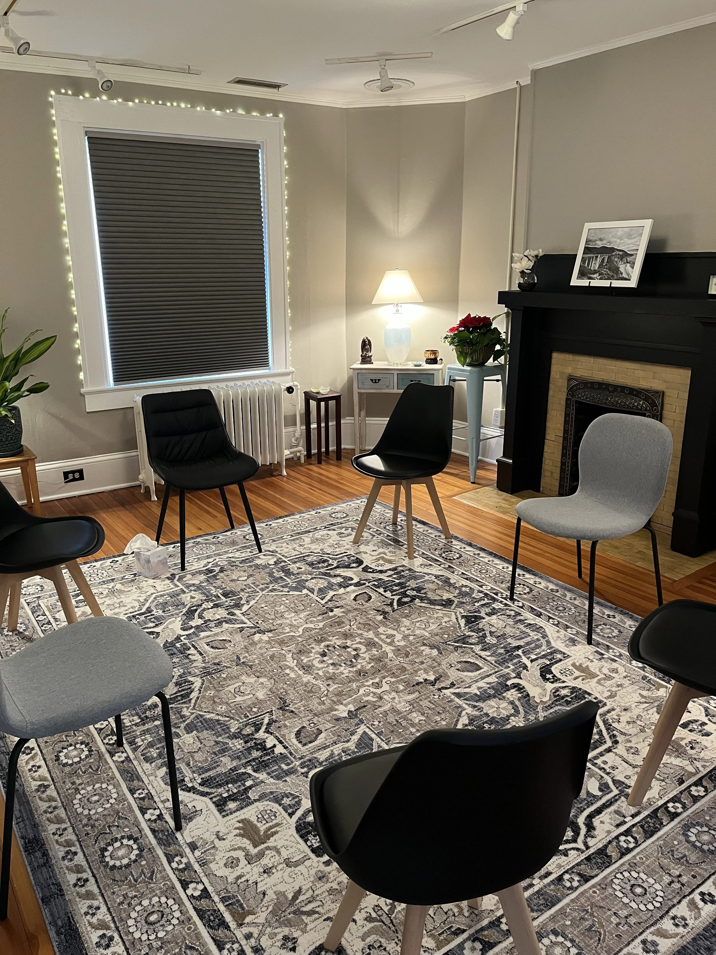 A cozy living room with yellow walls, carpeted floor, and seven chairs of various styles and colors arranged around the room. There is a white door in the background, a wooden chest of drawers with a plant and decorative items on top, and a ceiling fan.