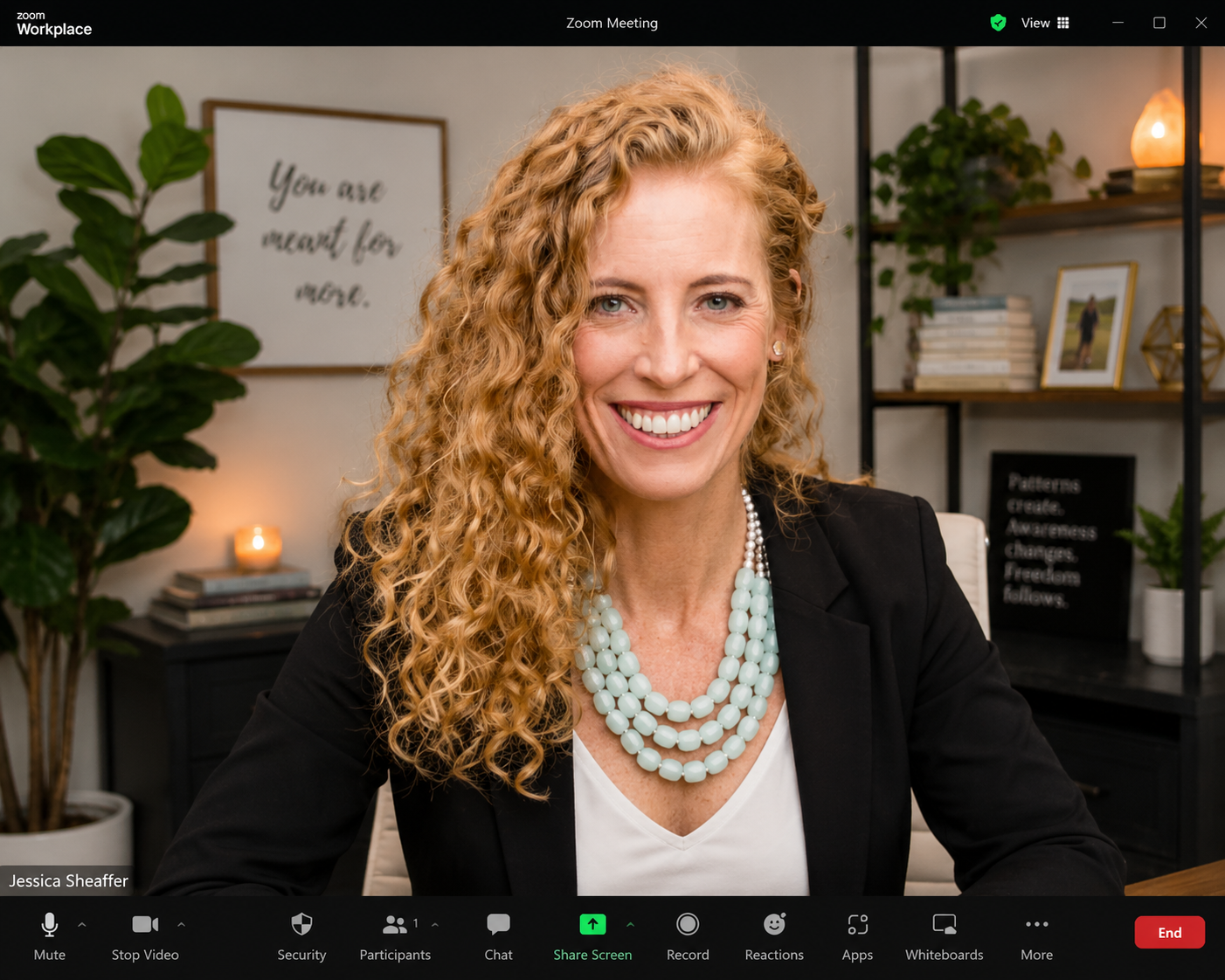 A woman with curly blonde hair smiling at the camera in a Zoom workplace meeting. She is wearing a black blazer and a multi-strand light blue beaded necklace. In the background, there is a large green plant, a framed quote that says 'You are meant for more,' a bookshelf with books, a framed photograph, a small lamp, and a black sign with the words 'Patterns create. Awareness. Changes. Freedom follows.'