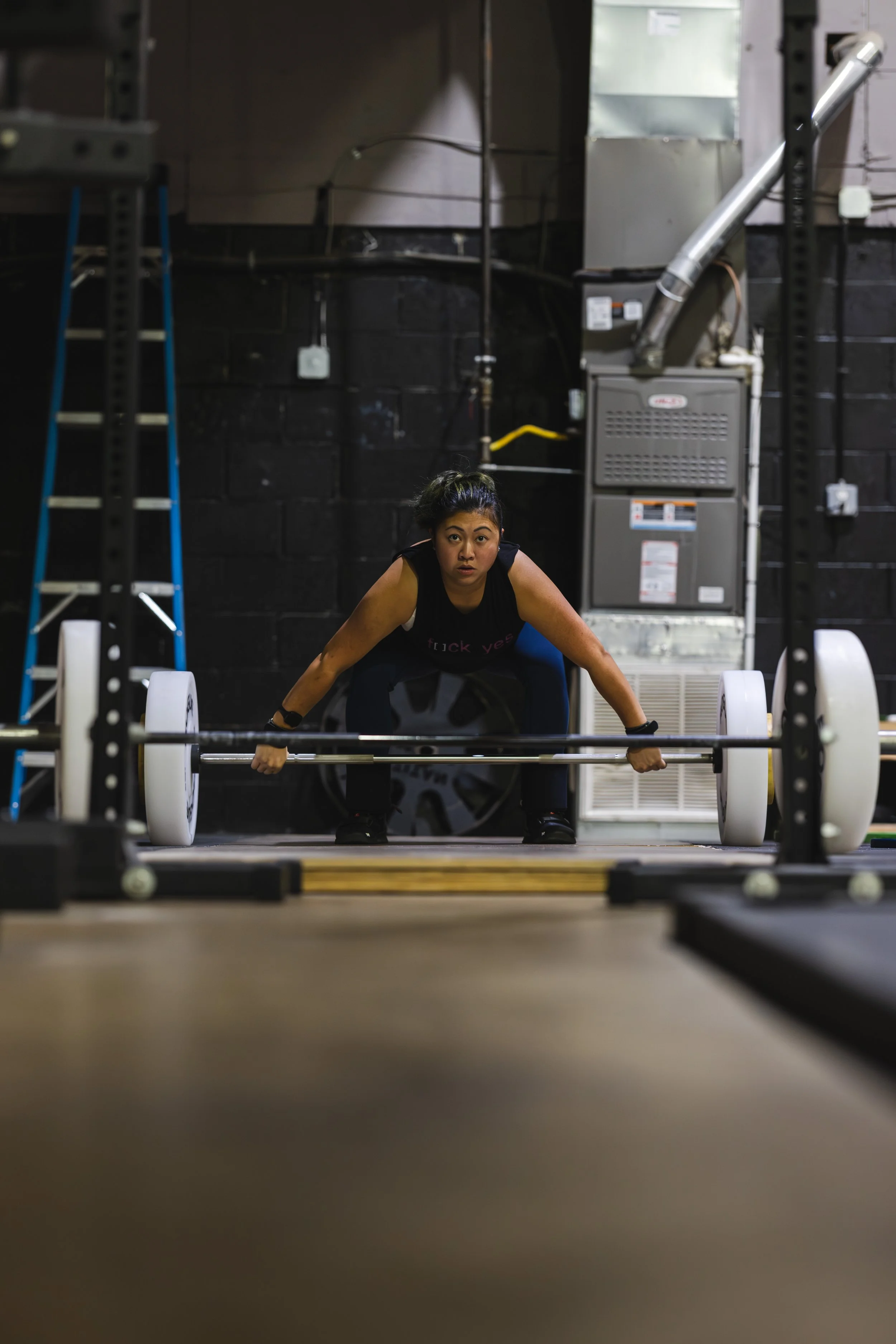 A woman prepares to lift a barbell in a gym, crouching down with a focused expression.