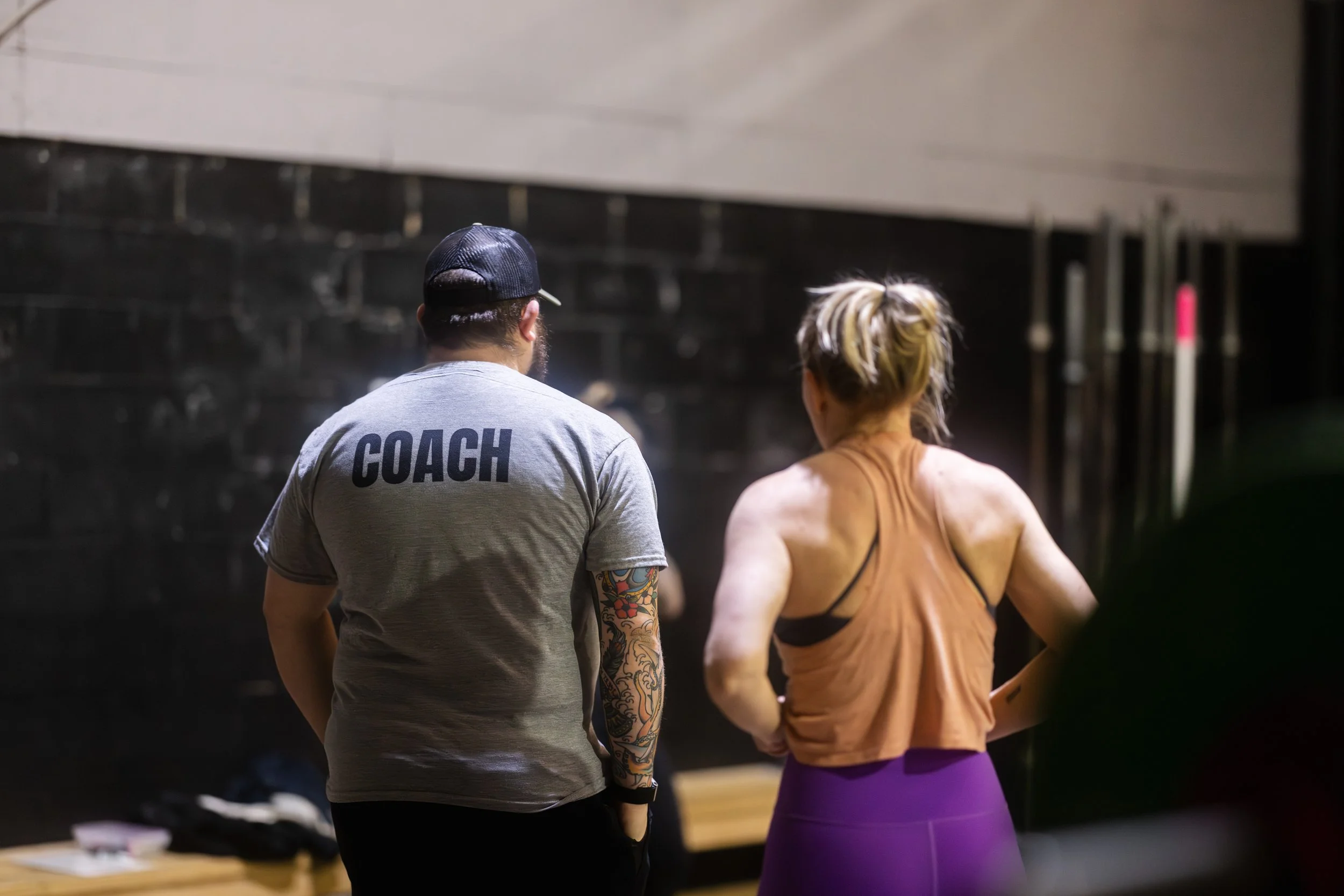 A man and woman stand together in a gym, with the man wearing a gray T-shirt labeled 'COACH' on the back, and the woman in a brown sleeveless top and purple leggings, both facing away from the camera.