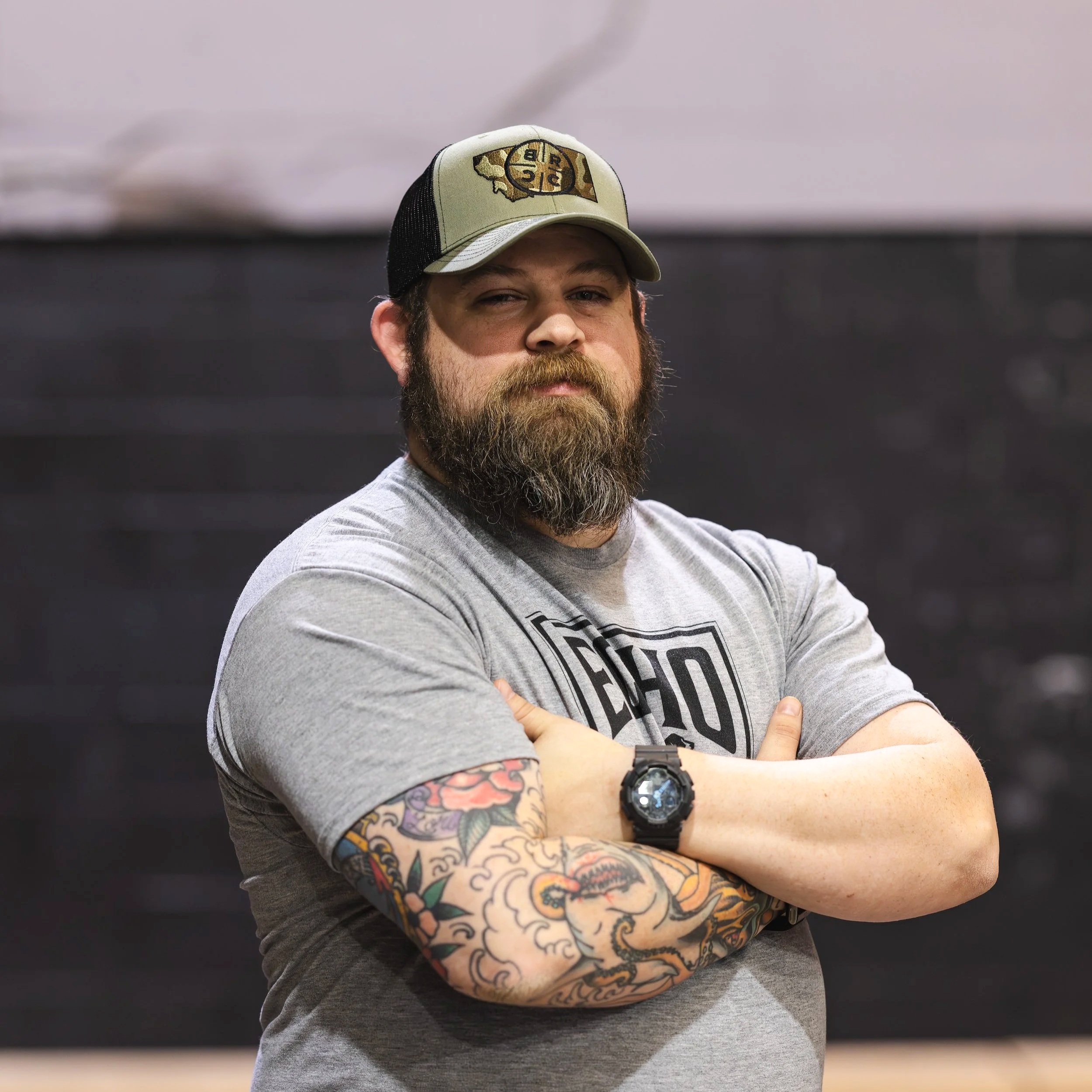 A man with a beard and tattoos on his left arm stands with his arms crossed, wearing a gray t-shirt, a black watch, and a baseball cap. The background is dark, and the man looks directly at the camera with a serious expression.